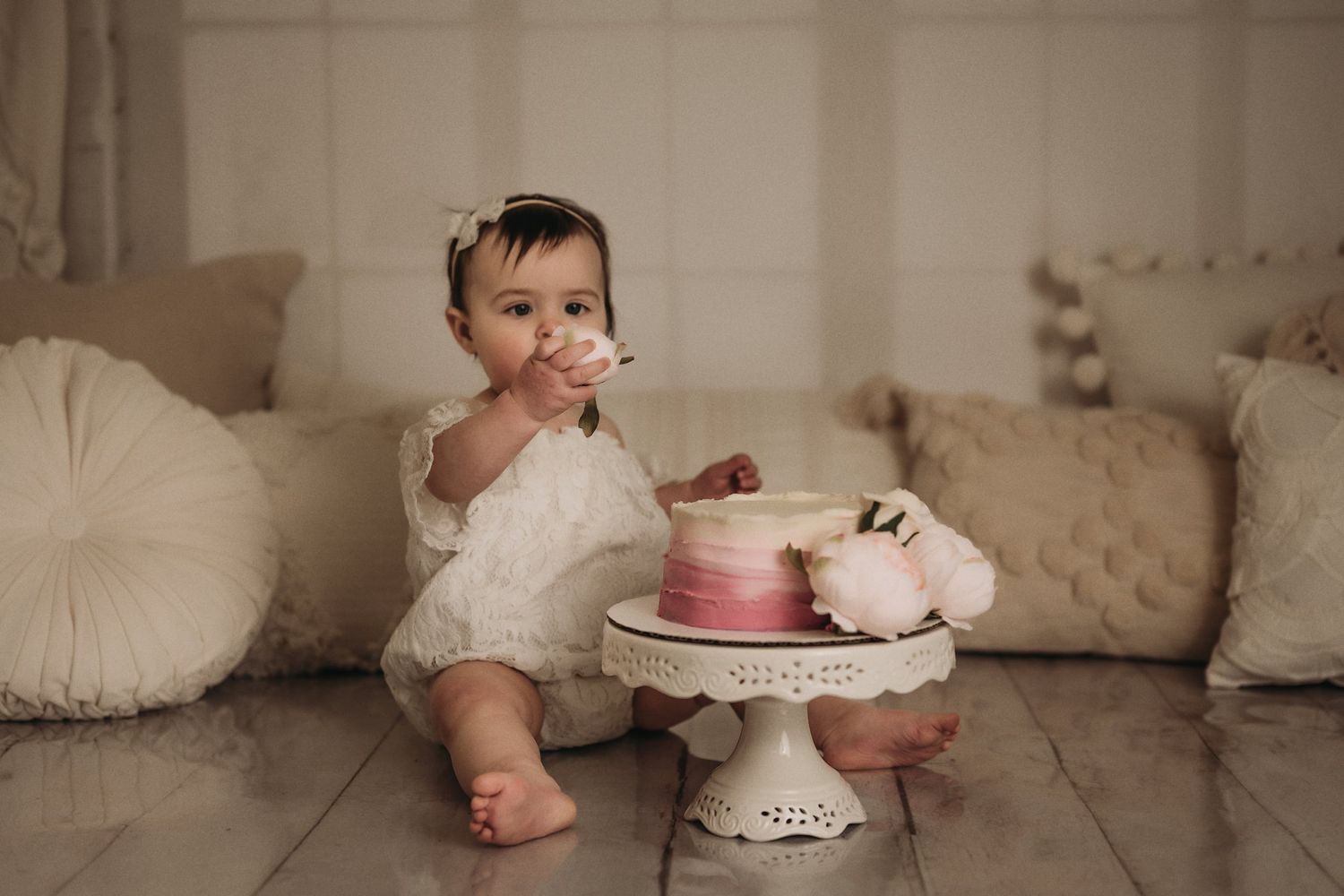 A small cake with pink ombre frosting and roses displayed on white pedestal stand with cozy decor.