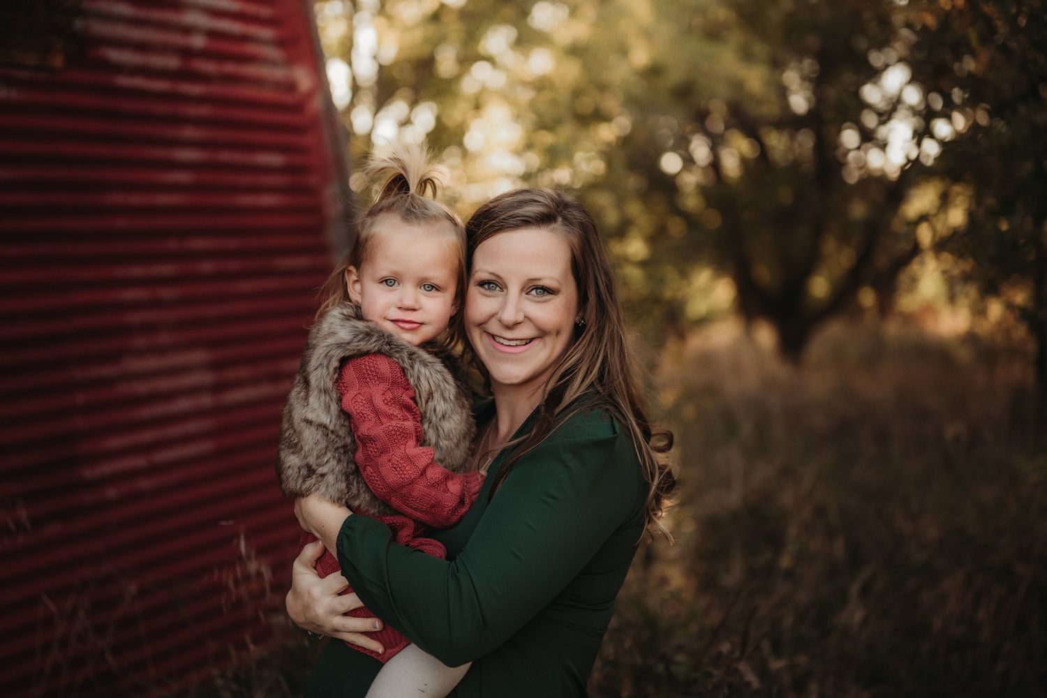 Tender moment captured between parent and child against red barn in golden light.