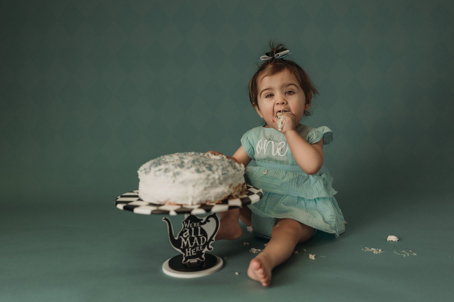 A first birthday celebration setup with a decorated cake on a black and white striped stand against a green backdrop.