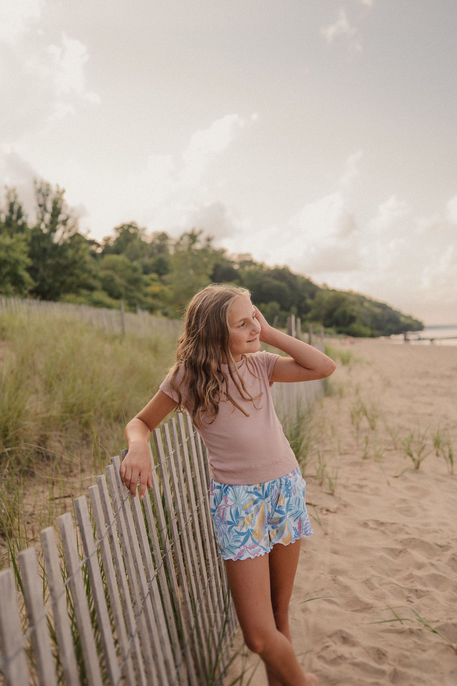 A person in pink top and patterned shorts leans against a beach fence during golden hour on a sandy beach.