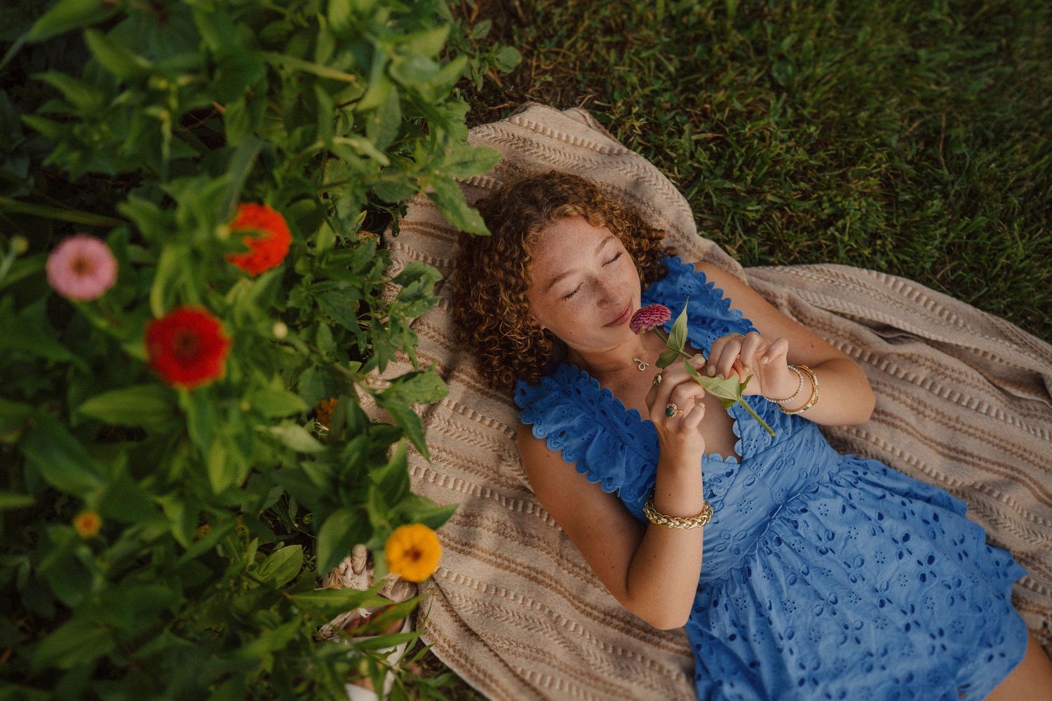 A person in a blue floral dress lies peacefully on a beige blanket among lush greenery and red flowers.