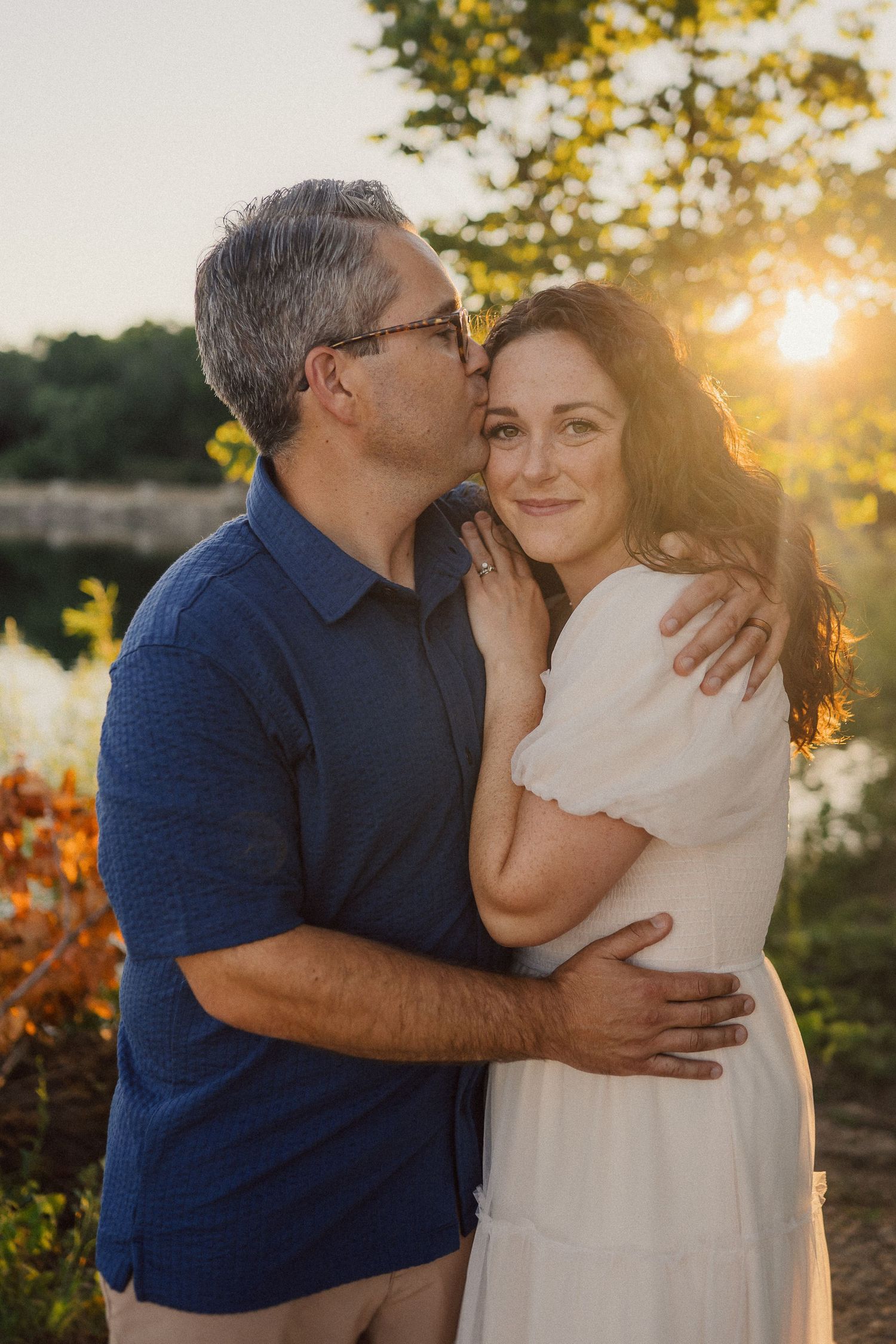Romantic couple shares intimate moment at sunset near water with fall colors surrounding them.