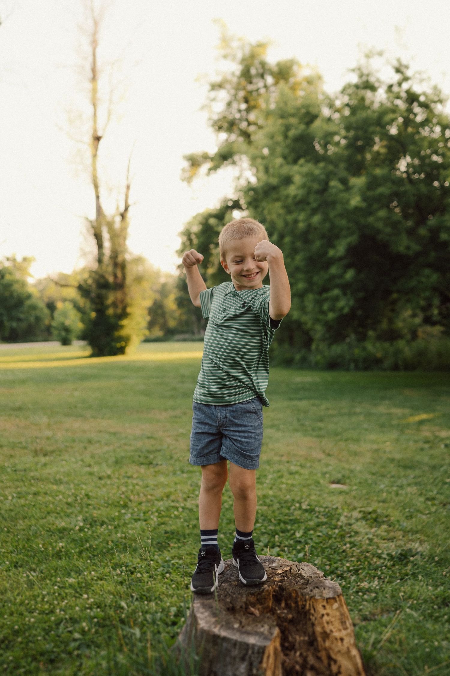 A young child stands on a tree stump in a green park during golden hour, wearing a t-shirt and shorts.