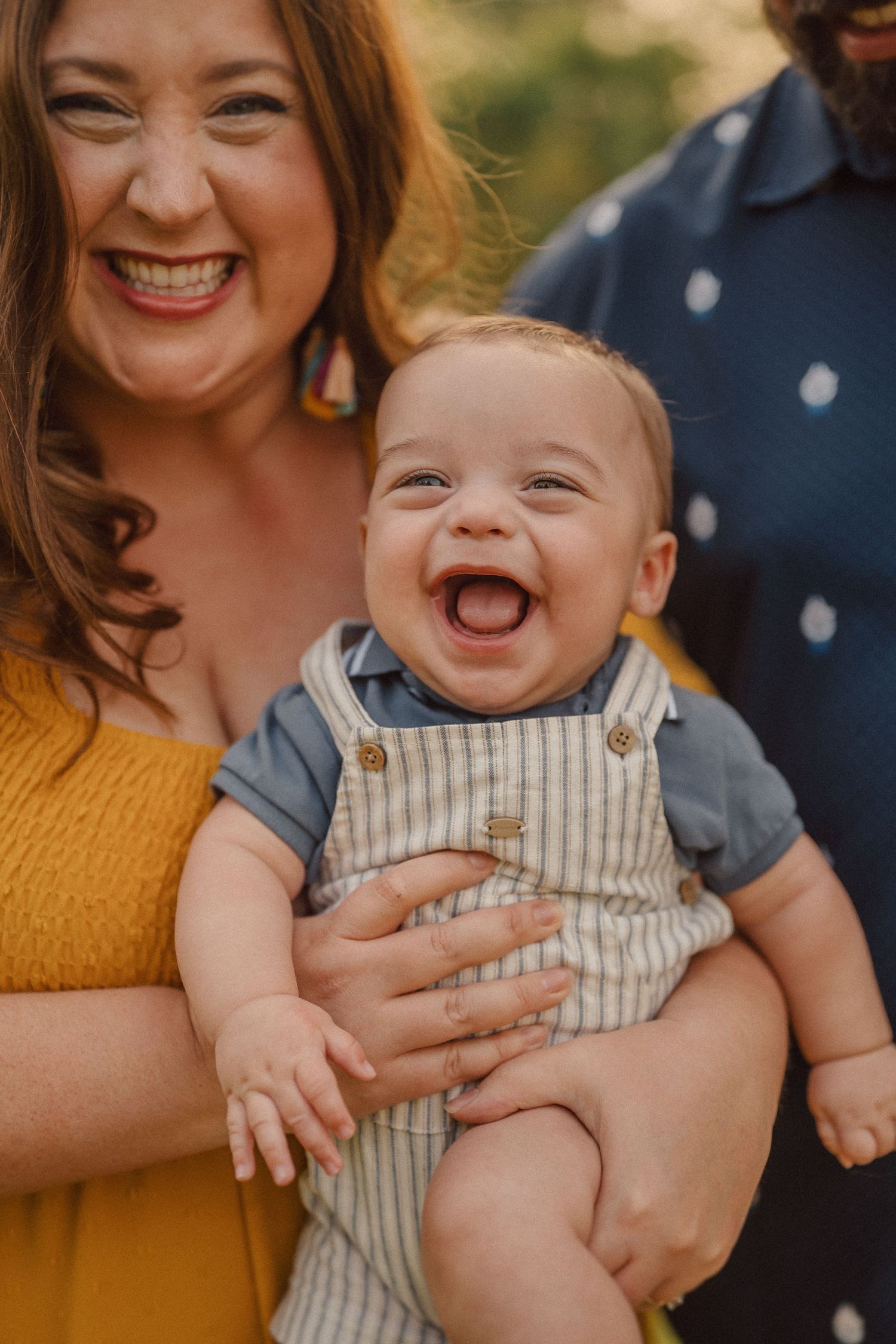 A baby in striped overalls laughs and smiles while being held in an outdoor photoshoot.