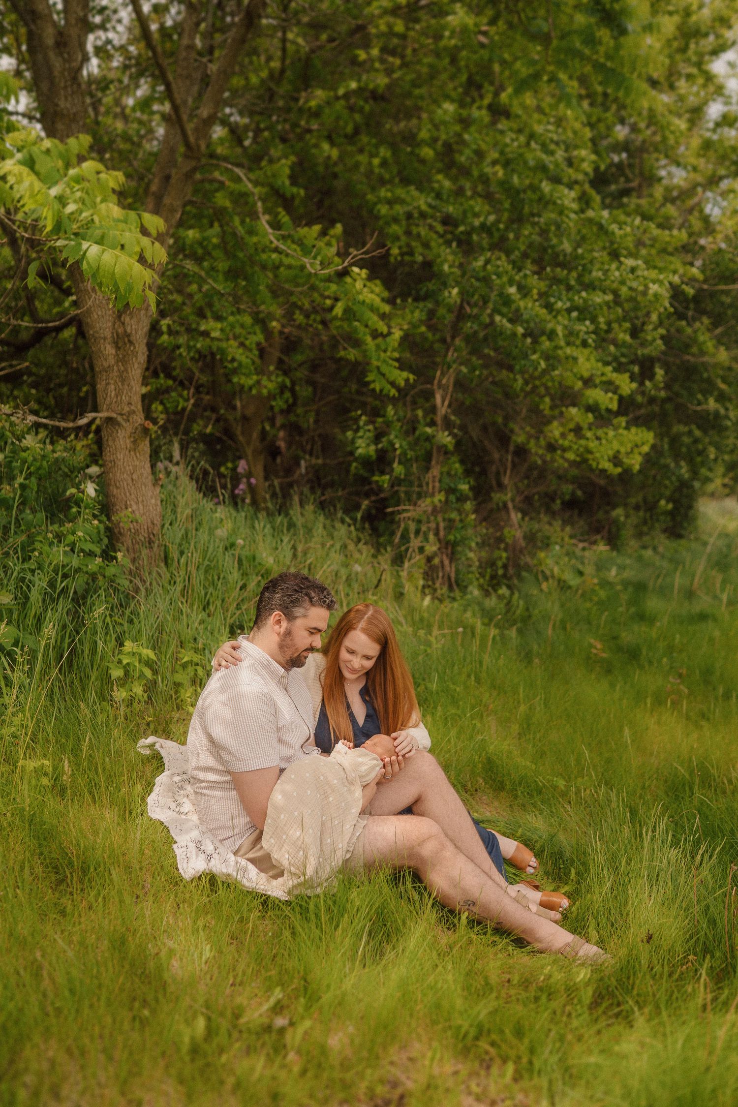 A couple sits together in tall summer grass near trees during a warm outdoor photo session.