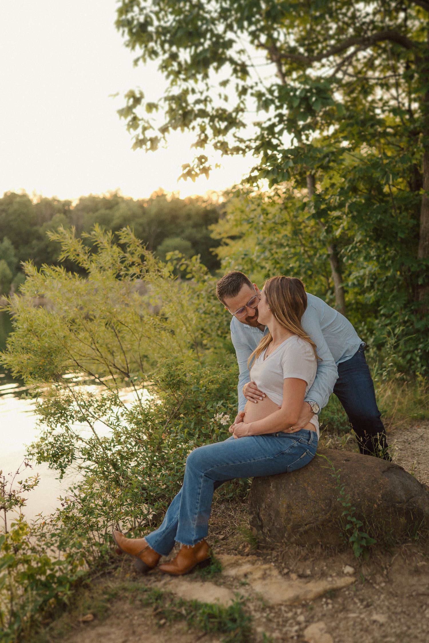 A romantic couple sits on a riverbank under trees during a golden sunset photoshoot.