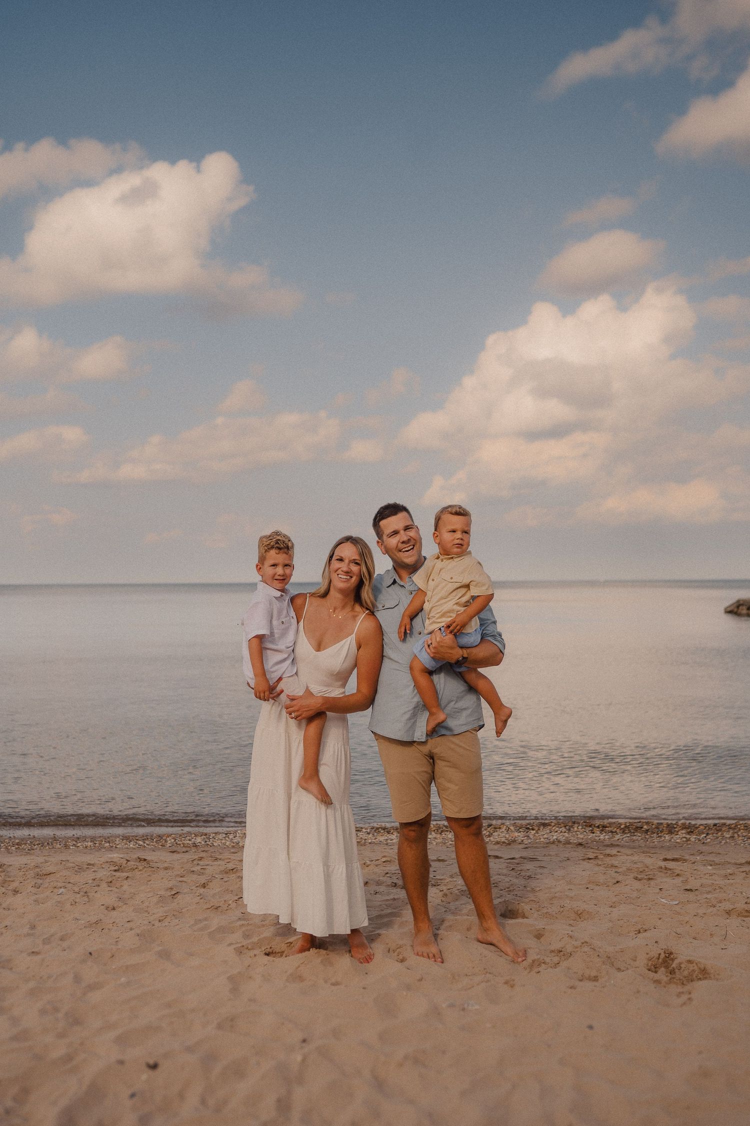 A family stands together on a sandy beach at sunset with fluffy clouds and calm waters in the background.