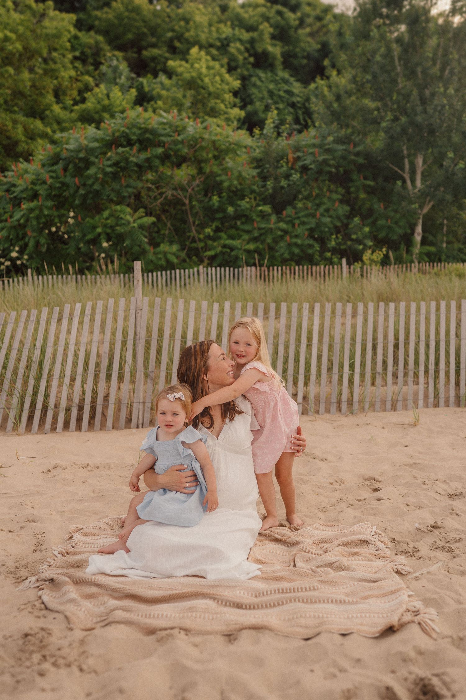 A family sits together on a knit blanket at the beach with wood fencing and green trees in the background.