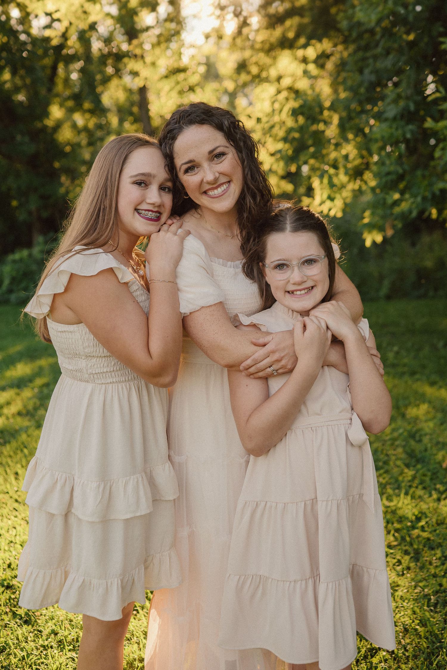 A family wearing coordinated cream-colored dresses embraces in a sun-drenched garden setting during golden hour.
