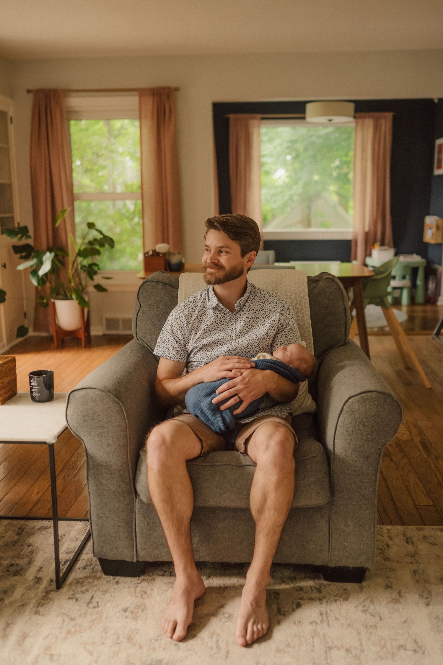 A relaxed person sitting barefoot in a grey armchair in a sunlit living room with plants.