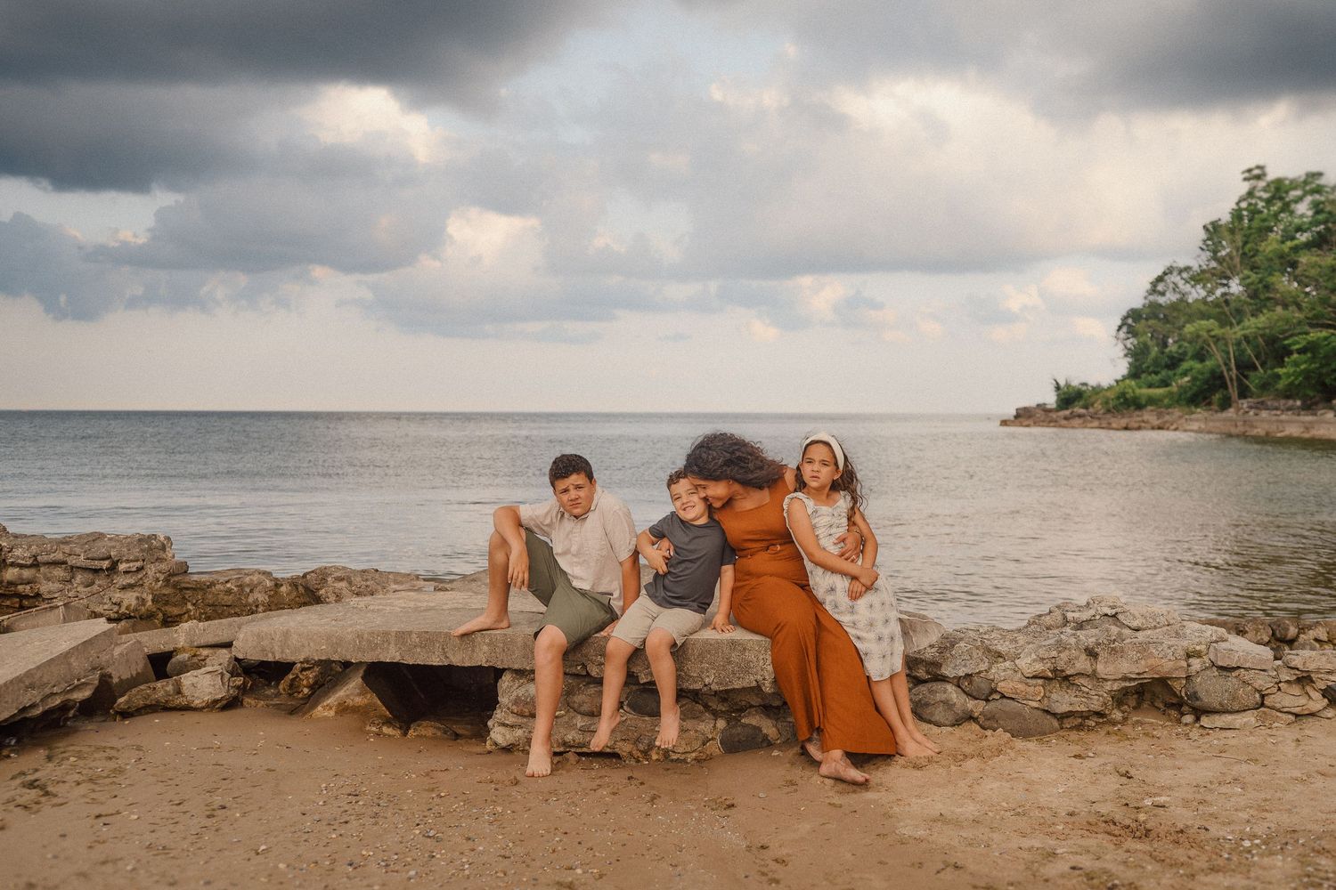 Three people sit together on coastal rocks at sunset with a moody cloudy sky and beach landscape in the background.