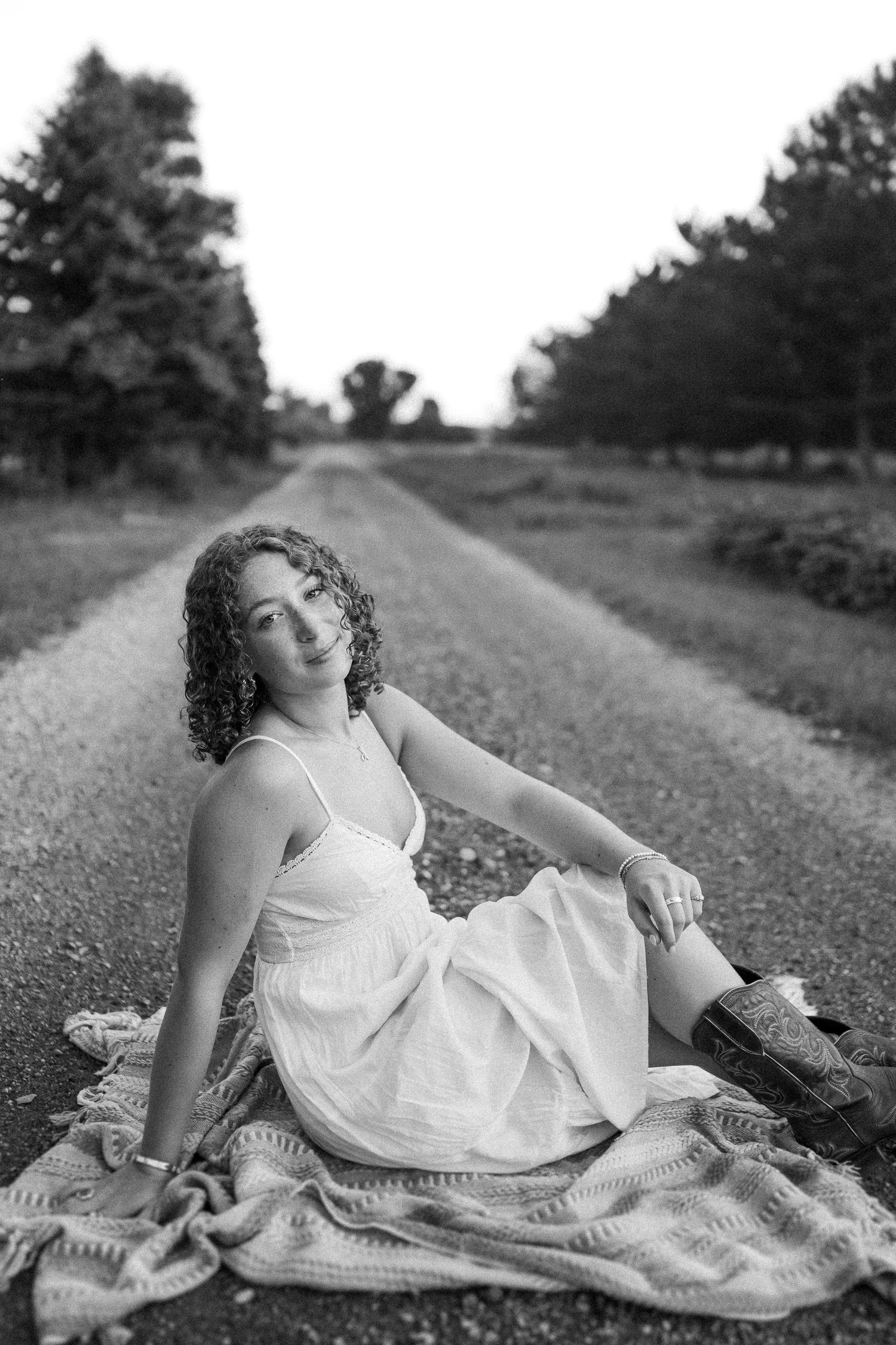 A person in a white dress reclines on a blanket along a rural country road in a black and white photograph.