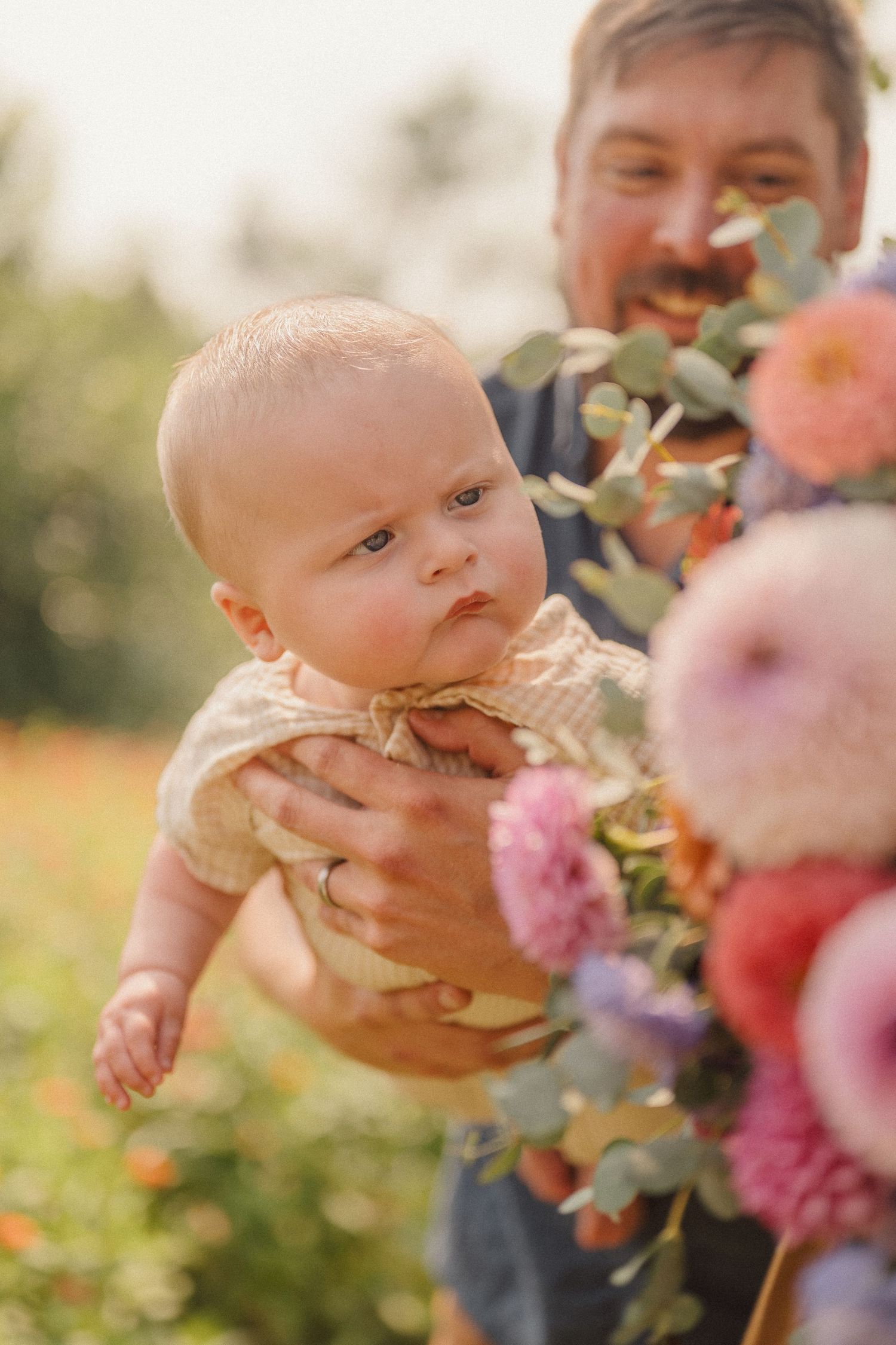Three playful photos of a baby and parent among pink flowers in a sunlit outdoor setting.