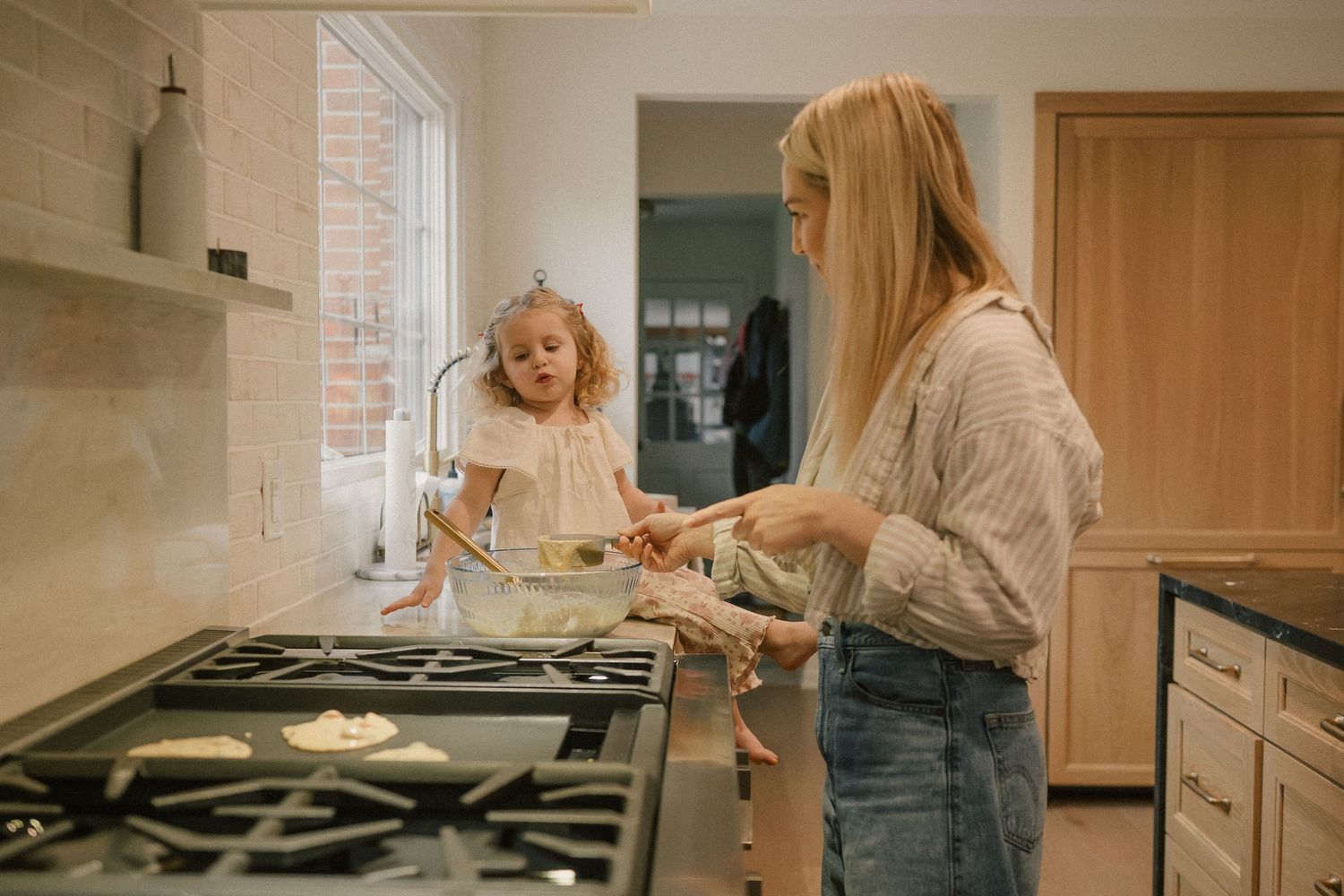 A blonde woman and young girl cooking pancakes together on a gas stove in a bright modern kitchen.