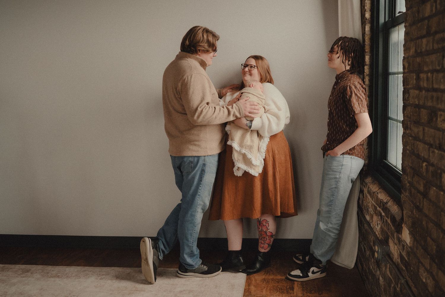 Three young adults in casual attire stand together near a window, sharing a warm moment indoors.