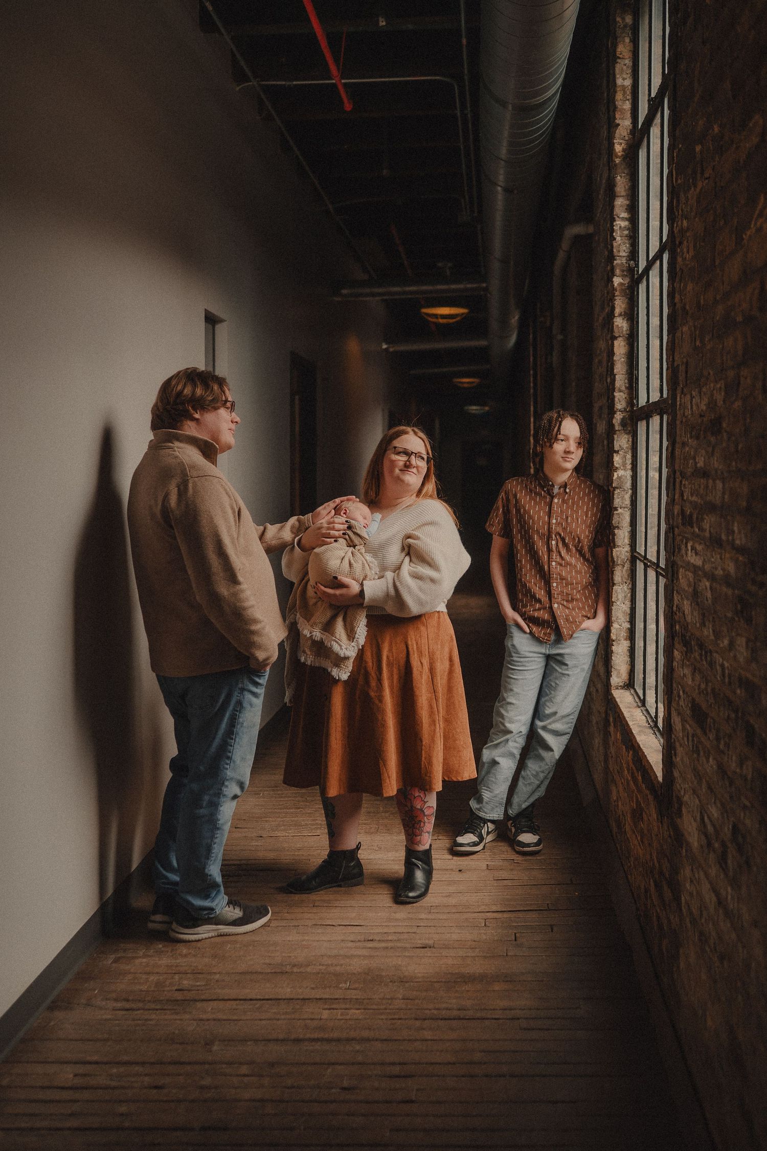 Three people standing in a rustic hallway with brick walls, one holding a baby, wearing casual earth-tone outfits.