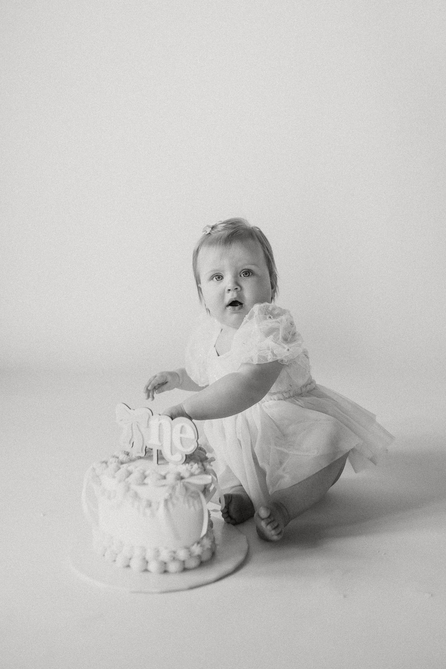 Black and white photo of a baby girl in a white dress at her first birthday cake smash.