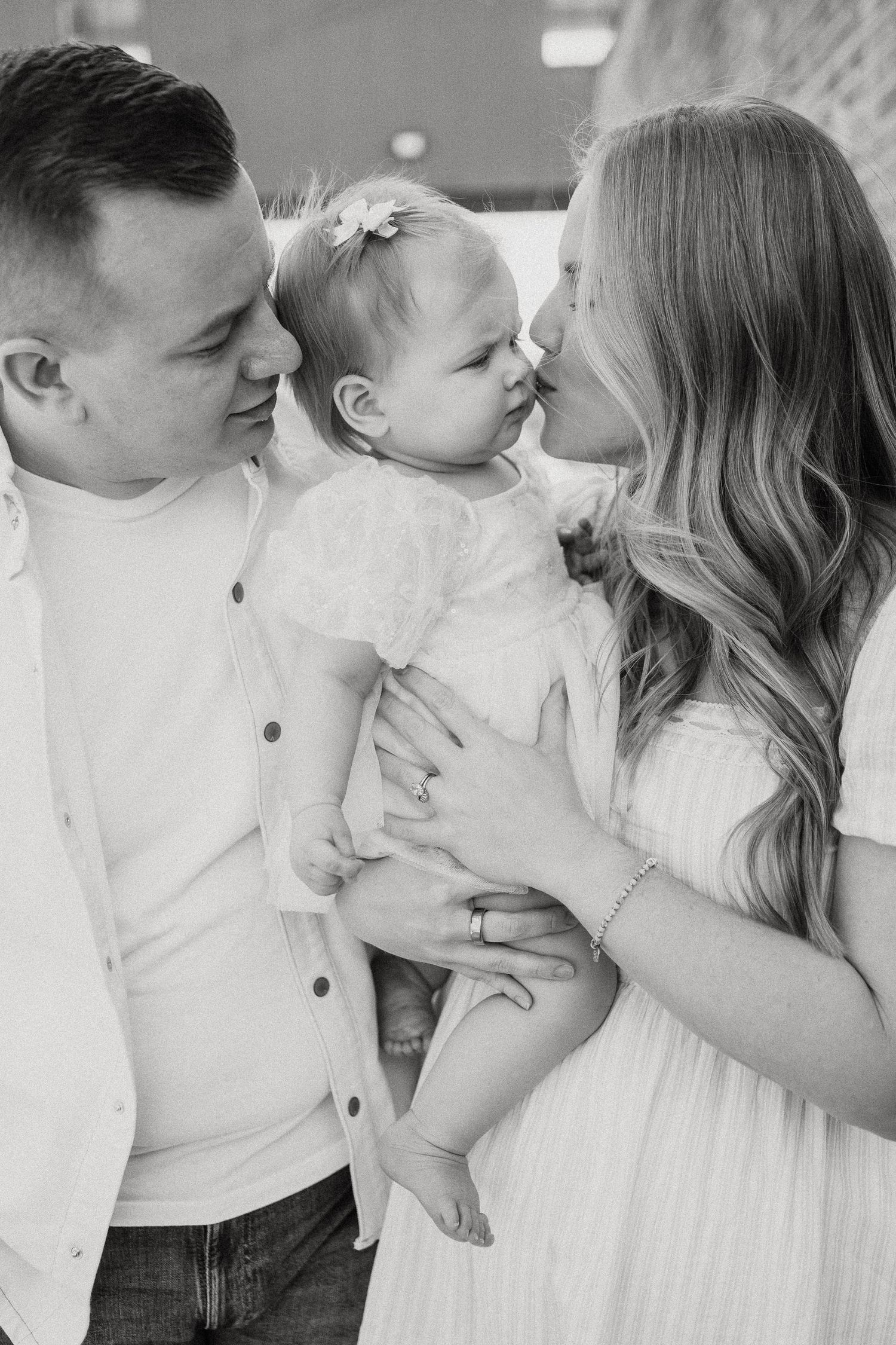 Black and white photo of young parents kissing their smiling baby girl in a white dress.