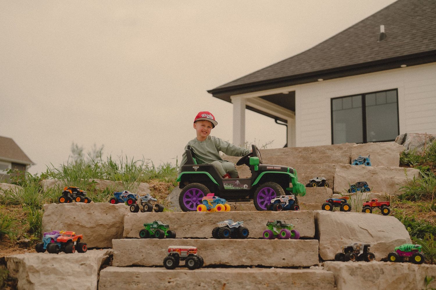 Boy in red cap sits on toy Jeep surrounded by monster truck collection on stone steps.