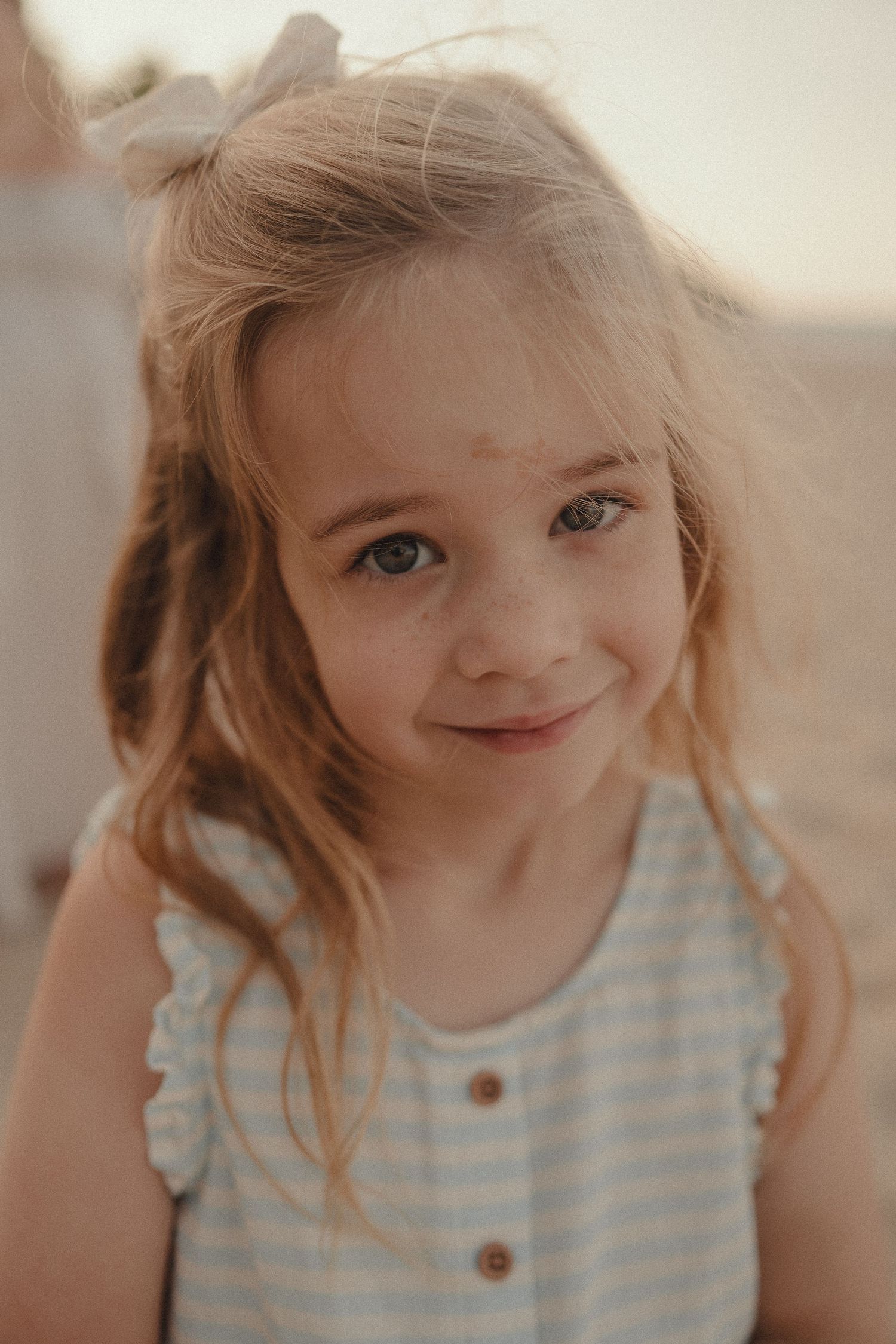 Close-up portrait of smiling young girl with freckles and bow in hair wearing striped dress outdoors.