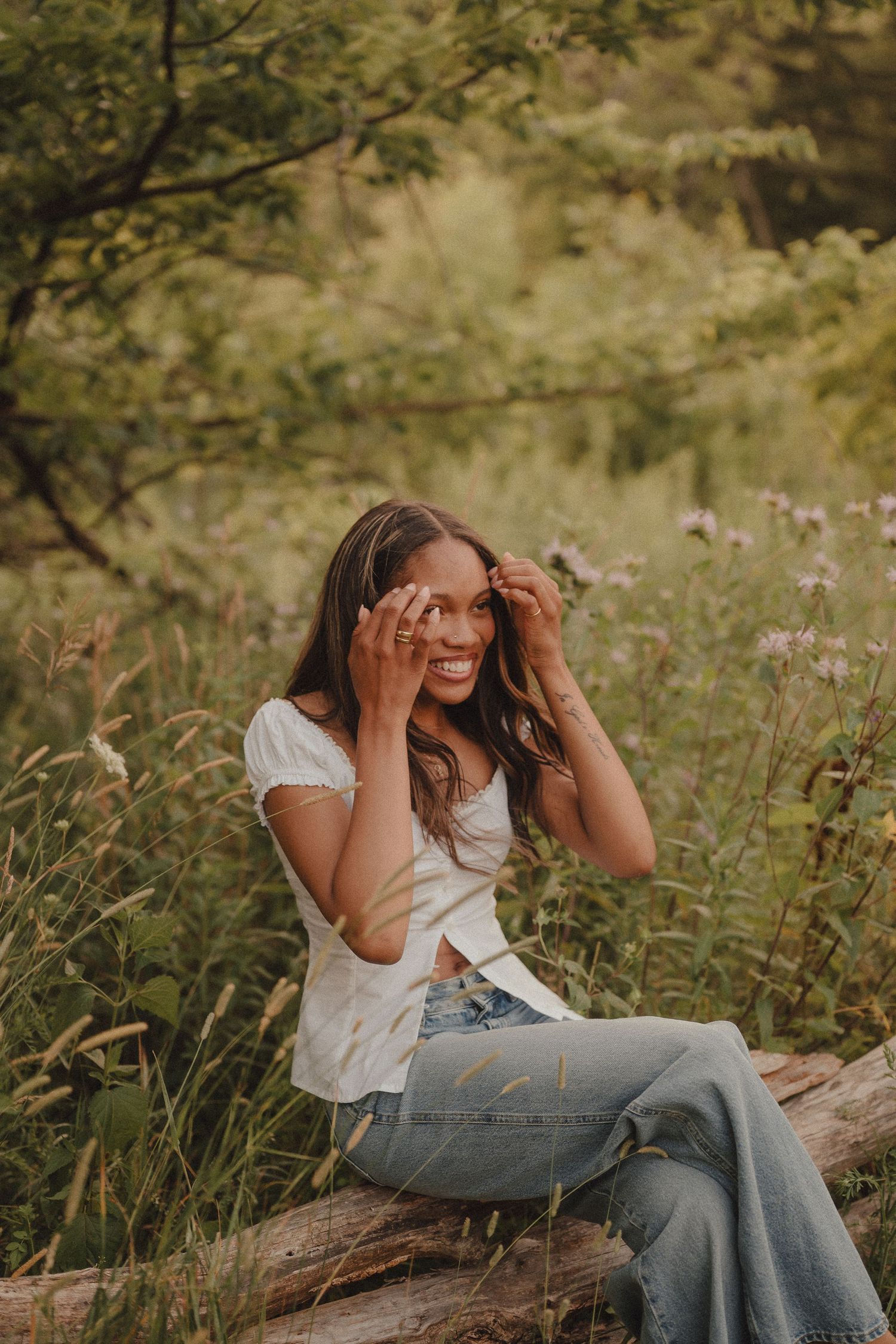 Young woman in white top and jeans sits on a log laughing in a lush green wildflower meadow.