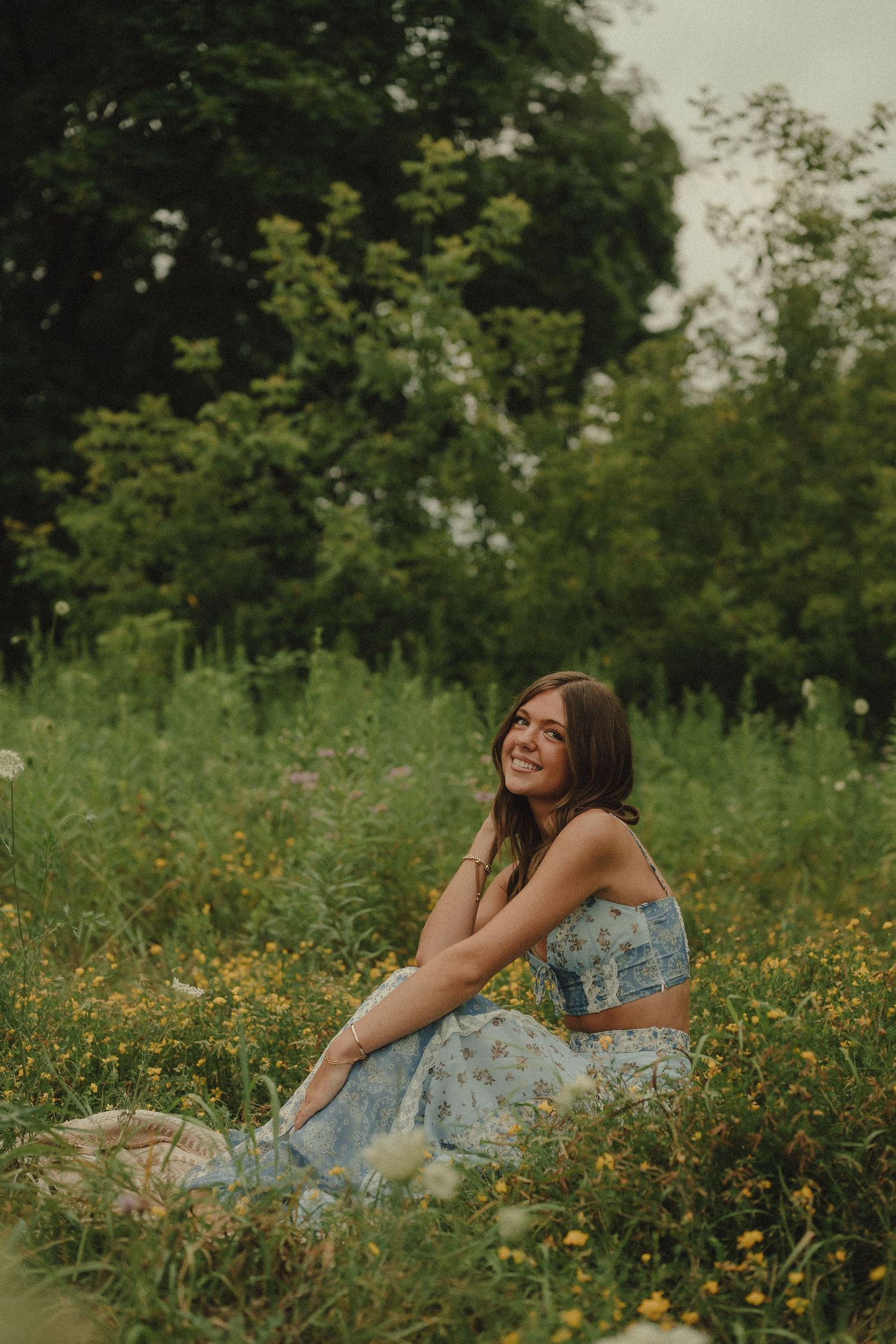 Smiling young woman in blue floral outfit sitting in colorful wildflower meadow with green trees.