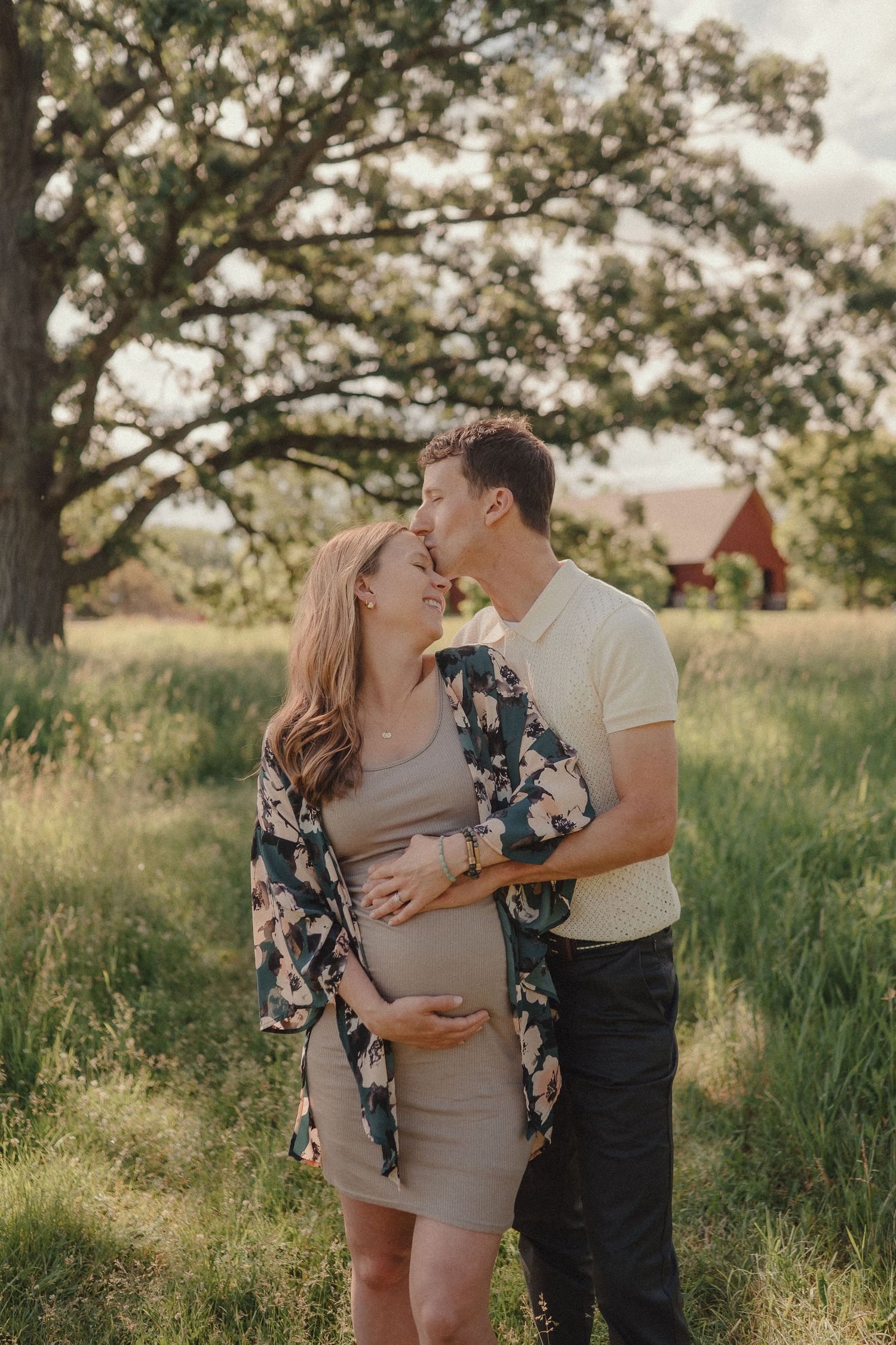A couple shares a tender kiss in a sunlit field, the woman cradling her pregnant belly near a red barn.
