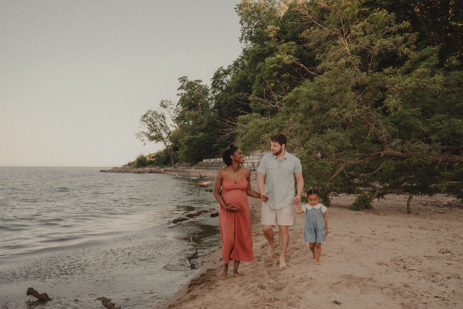 Pregnant woman walks with partner and child on sandy beach near lush trees at sunset.