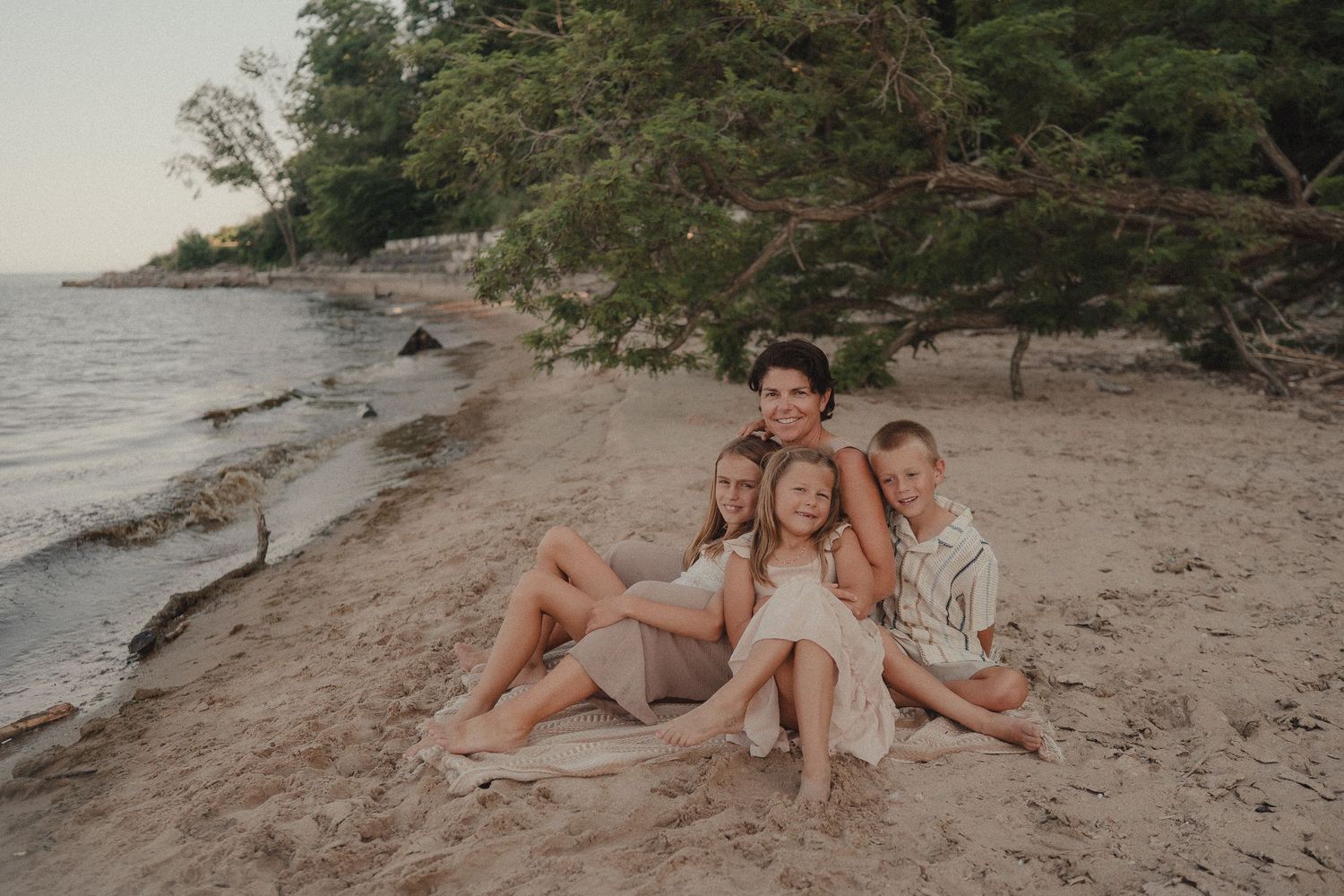 Family of four posing together on a sandy beach shore with trees and calm water in the background.