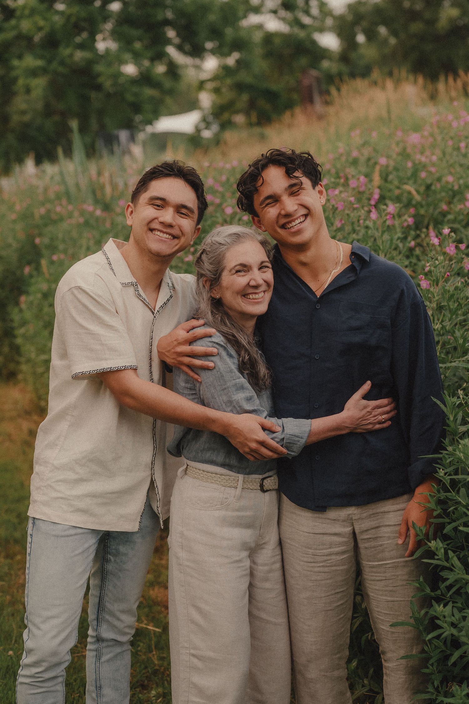 A smiling older woman is embraced by two young men in a colorful wildflower field outdoors.