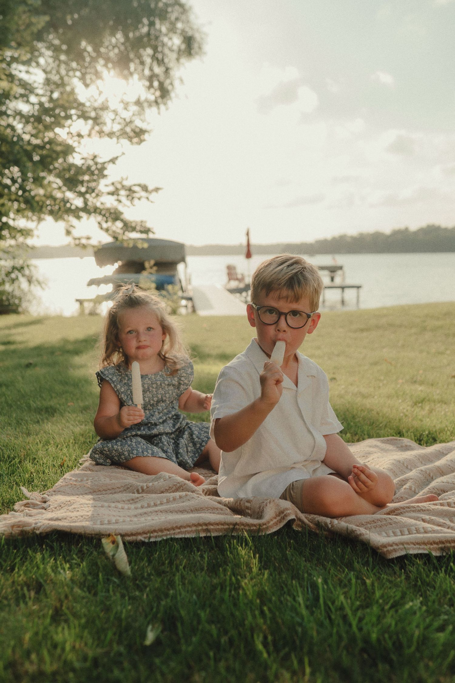 Two children sit on a blanket by a lake eating popsicles at golden hour.