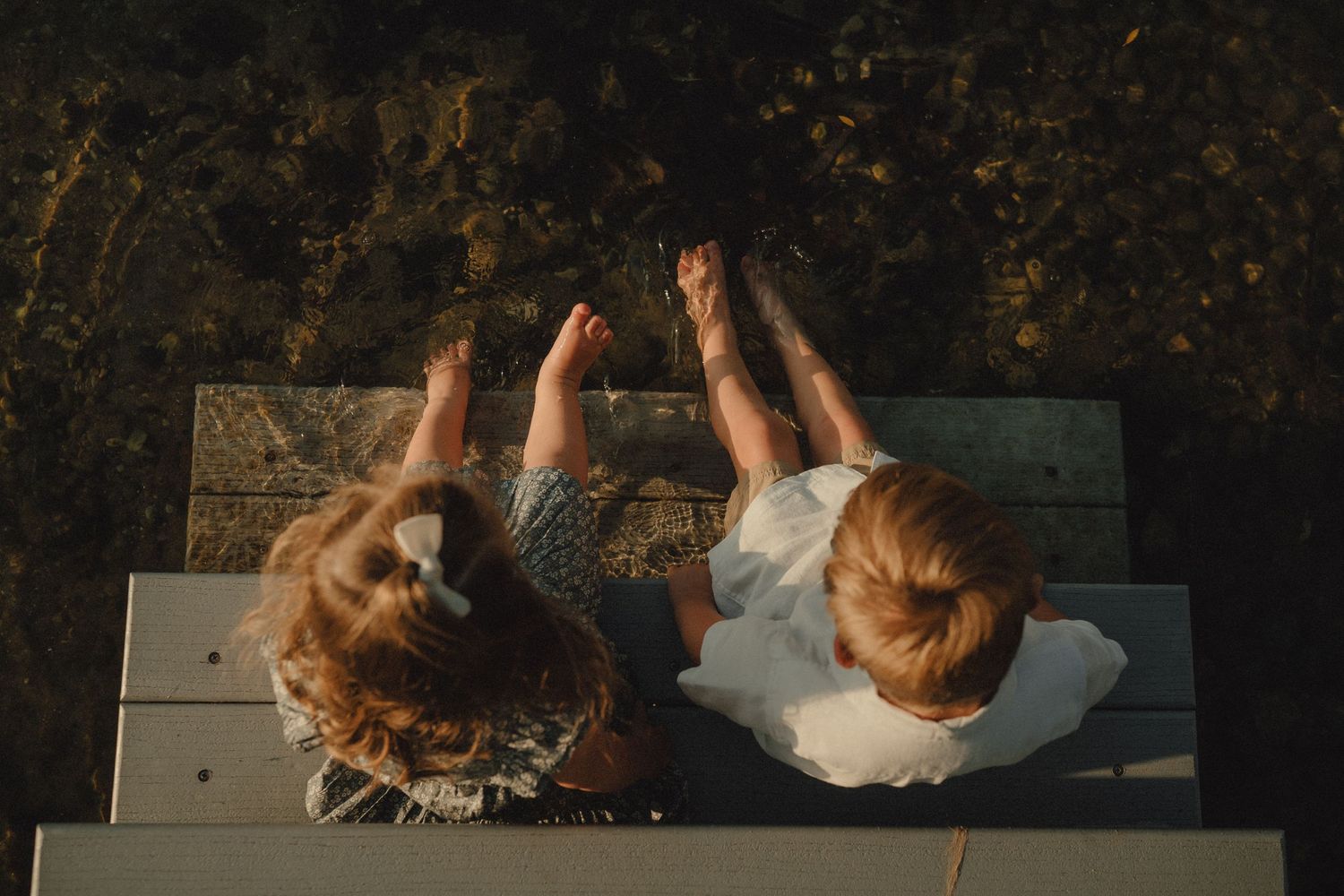 Overhead view of two young children sitting on a dock with their bare feet dangling over water.