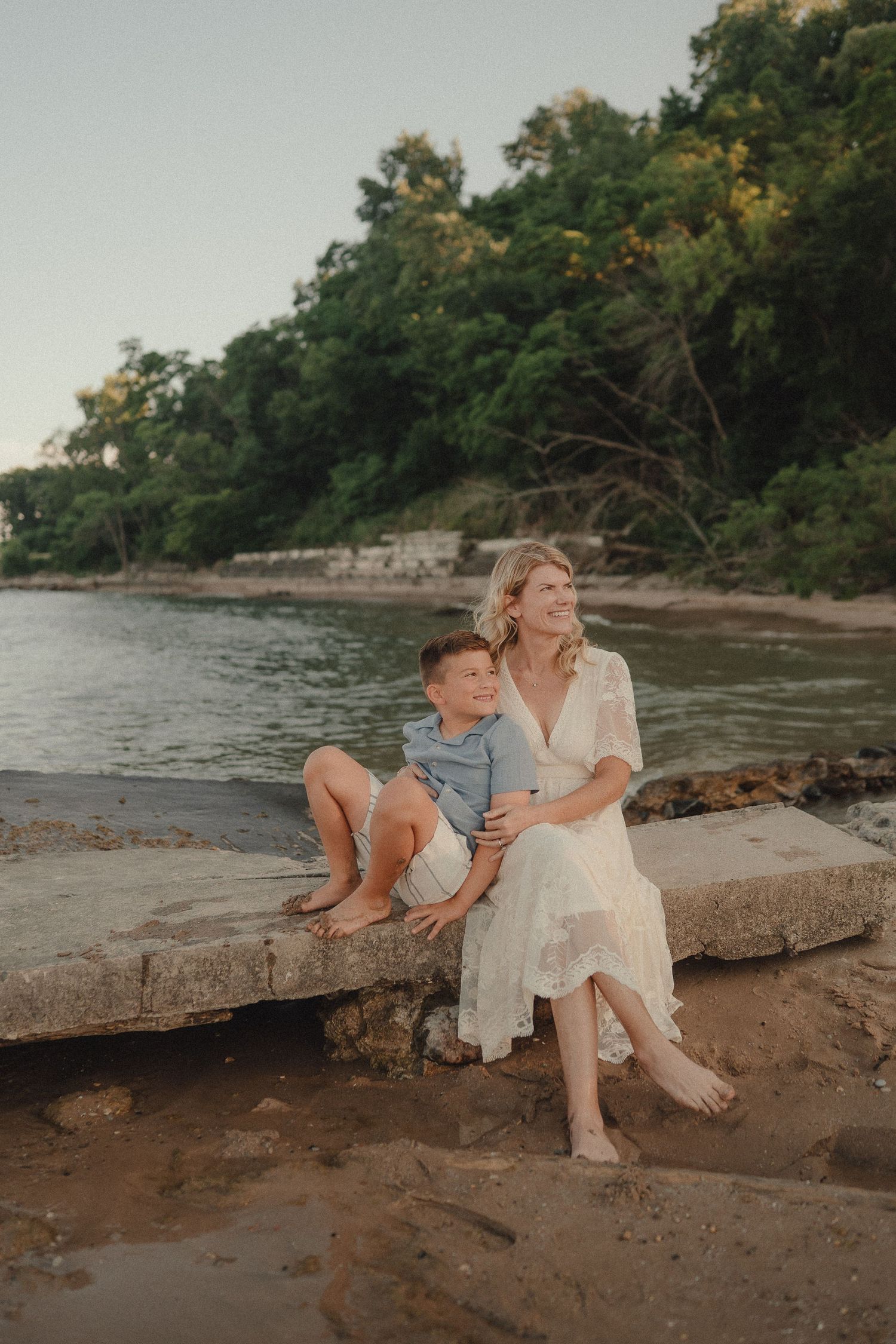 A woman in a white lace dress sits with a young boy on a rocky shore near calm water and lush green trees.