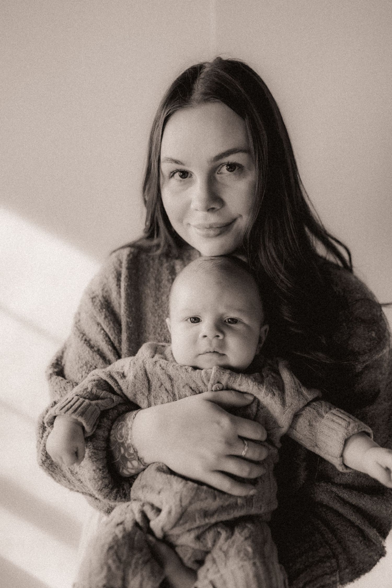 A black and white portrait of a parent holding an infant in a warm sweater with natural window light.