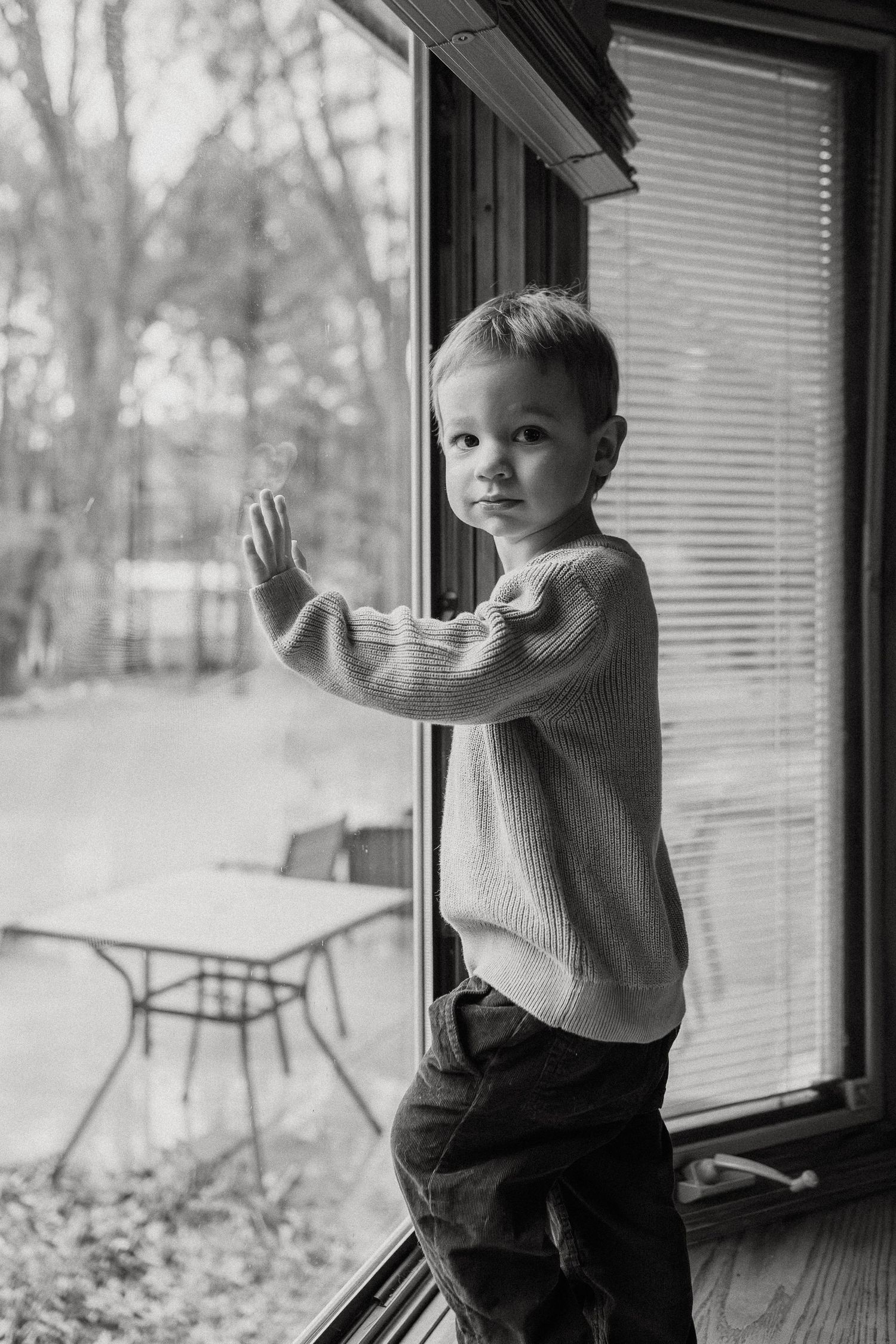 A toddler in a gray sweater plays and peeks through large windows with snowy scenery outside.