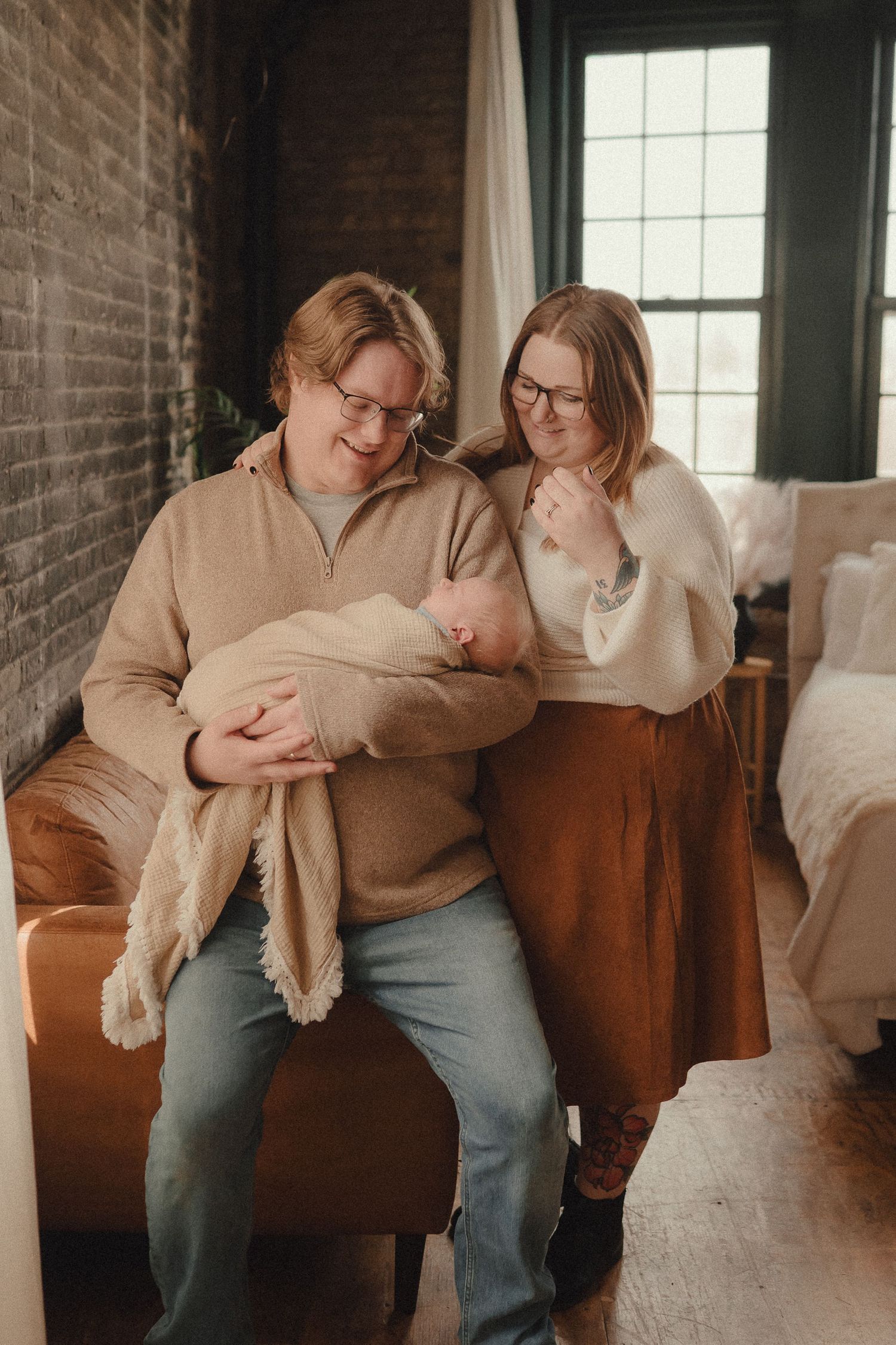 New parents sitting together, father holding swaddled newborn baby, mother leaning in close, in a cozy warmly lit room.