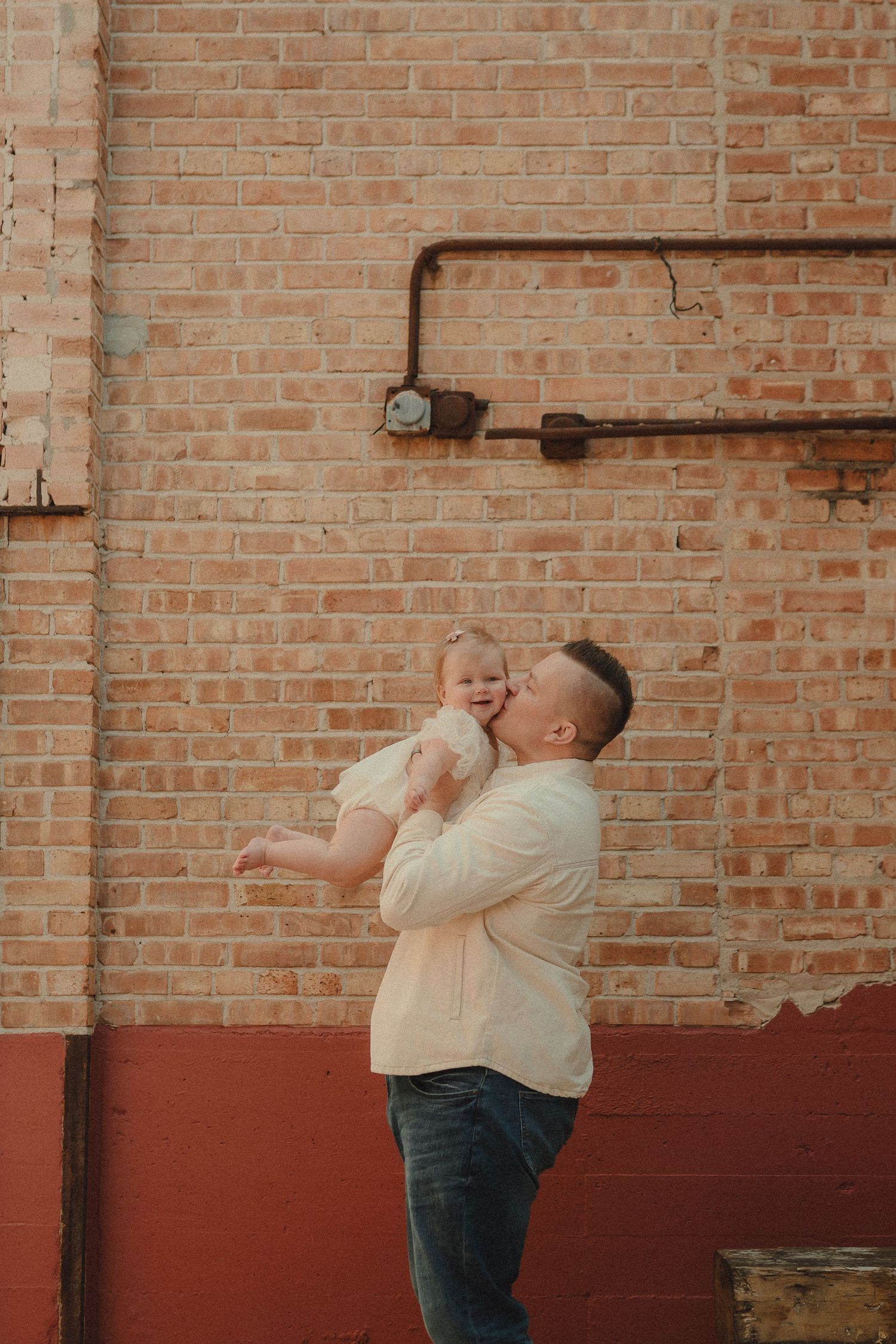 A father lovingly kisses his smiling baby girl while holding her up against a rustic brick wall backdrop.