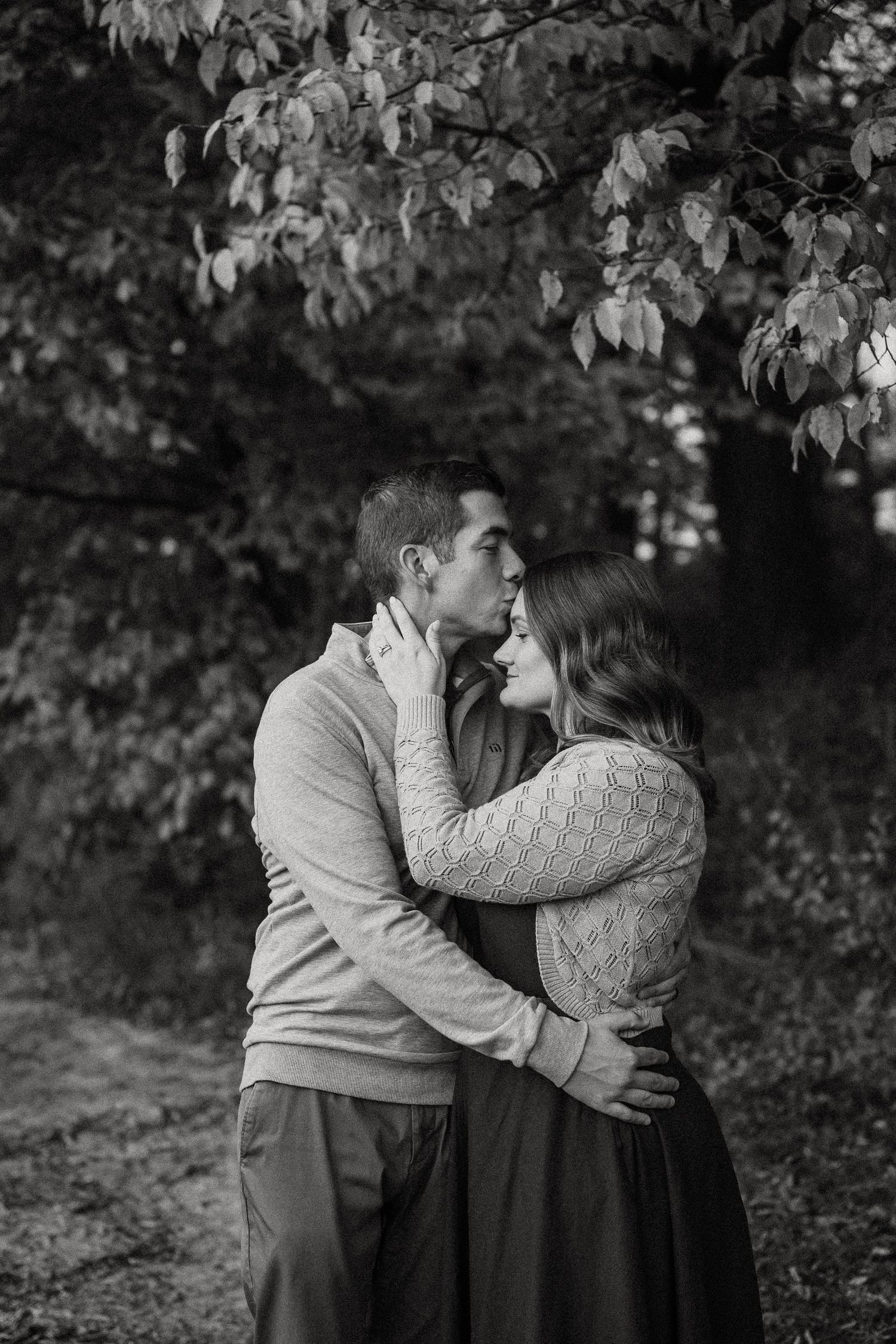 A romantic black and white portrait of a couple embracing under tree branches in a natural setting.