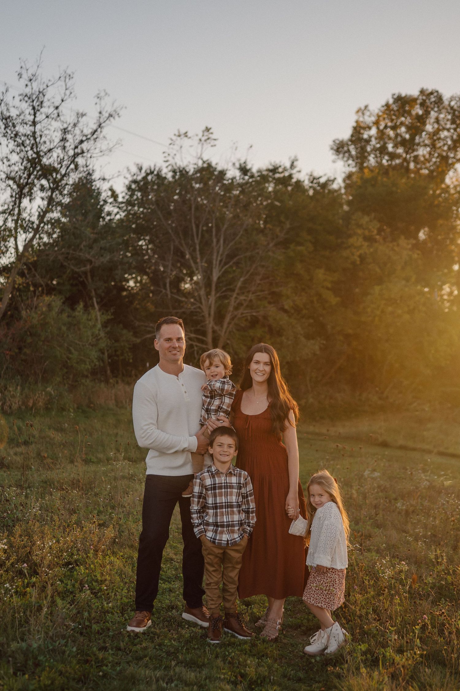 A family stands together in a golden field at sunset wearing coordinated autumn outfits.
