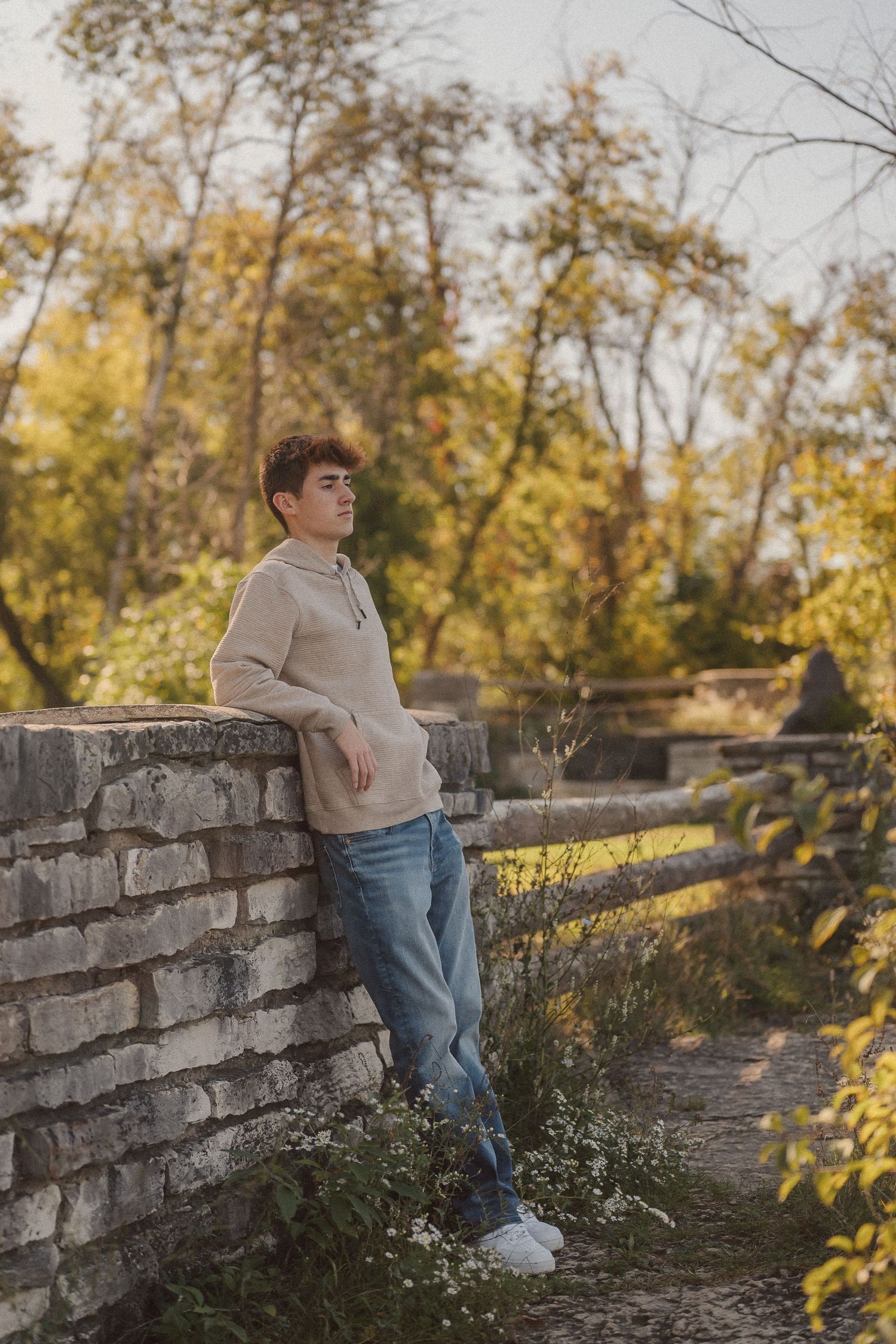Person in casual attire standing by a stone wall surrounded by fall foliage.