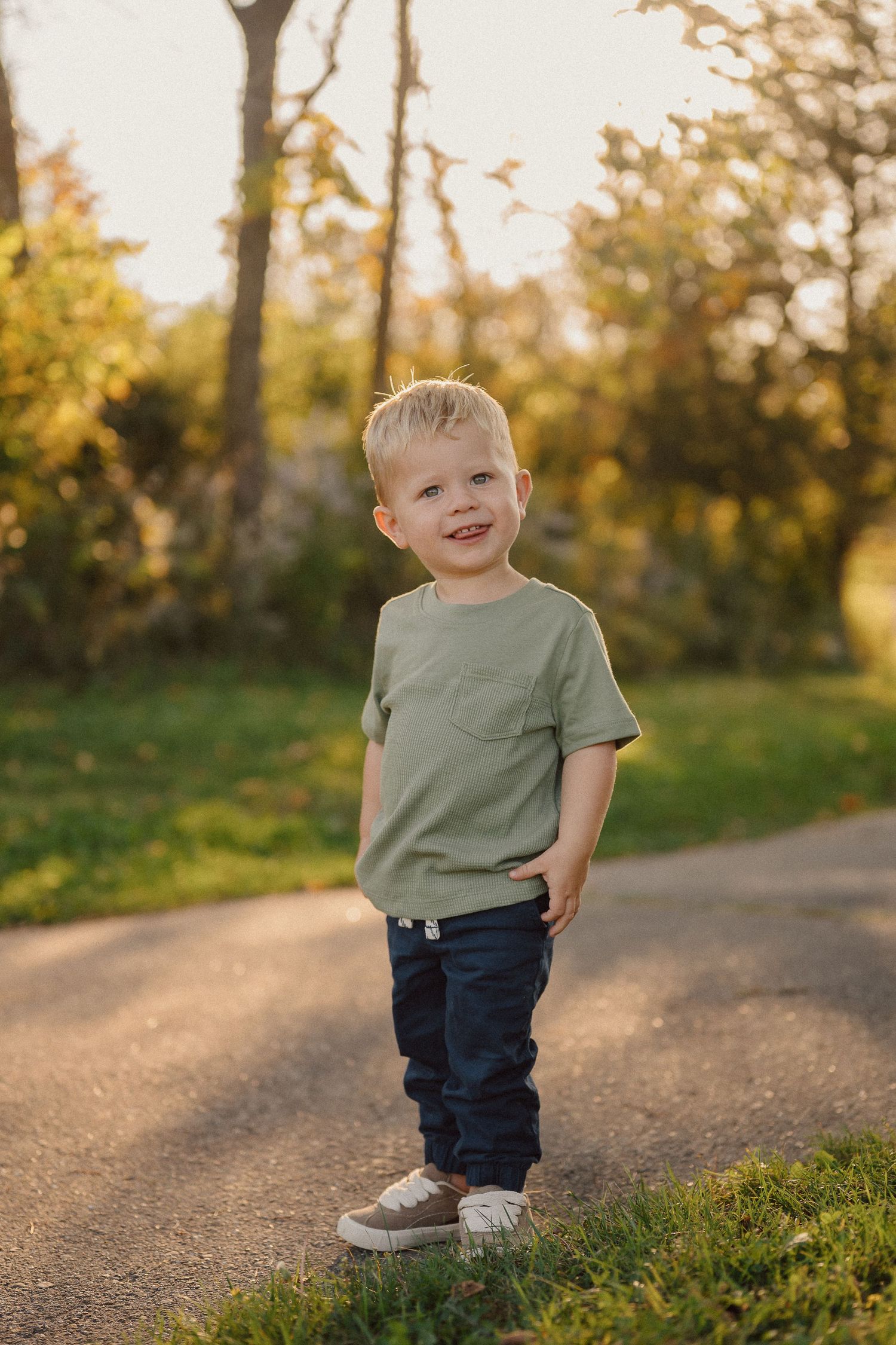 Young child in green shirt and jeans smiling while standing on outdoor path during golden hour.