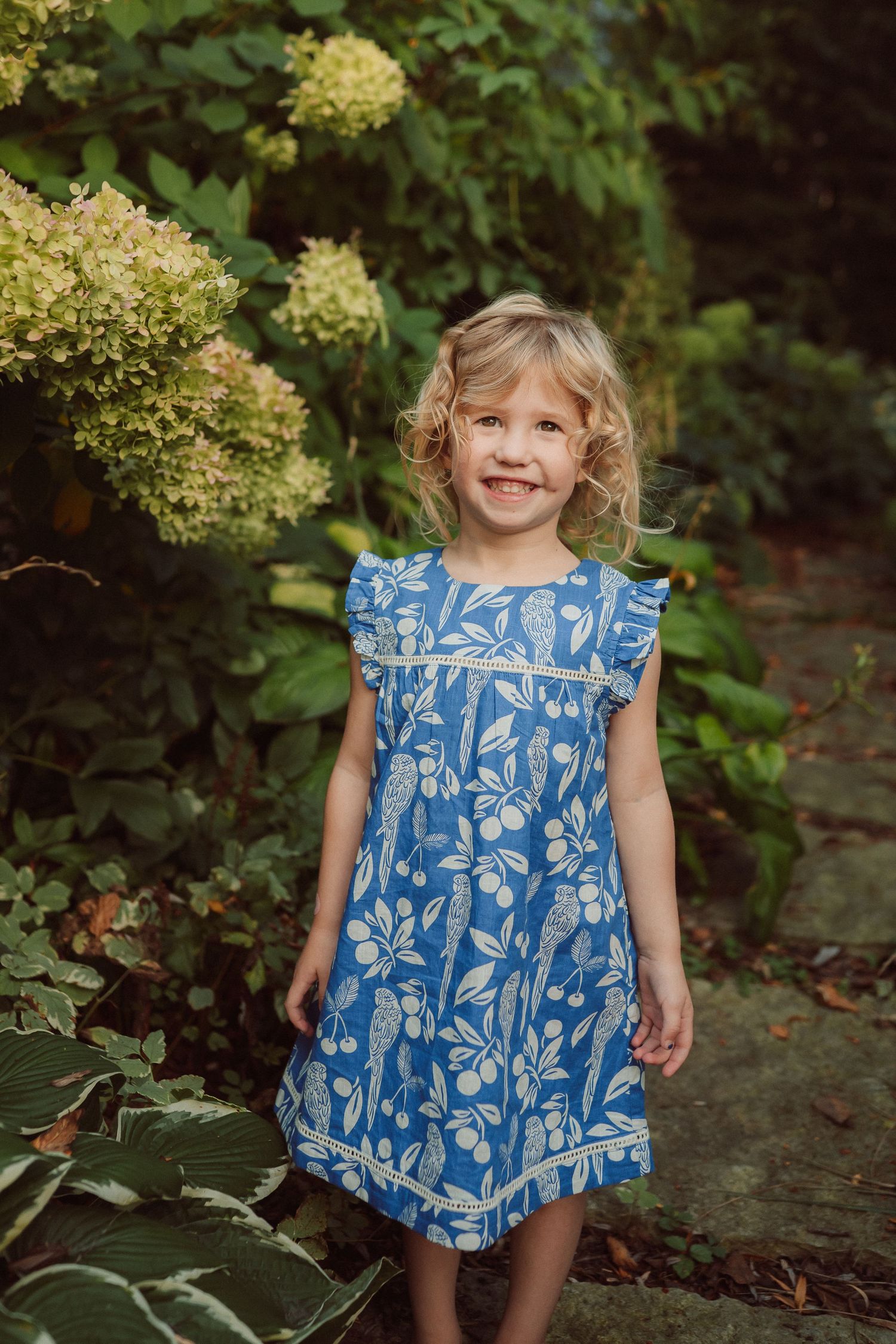 Young child wearing a blue and white floral dress poses playfully against a lush garden backdrop.