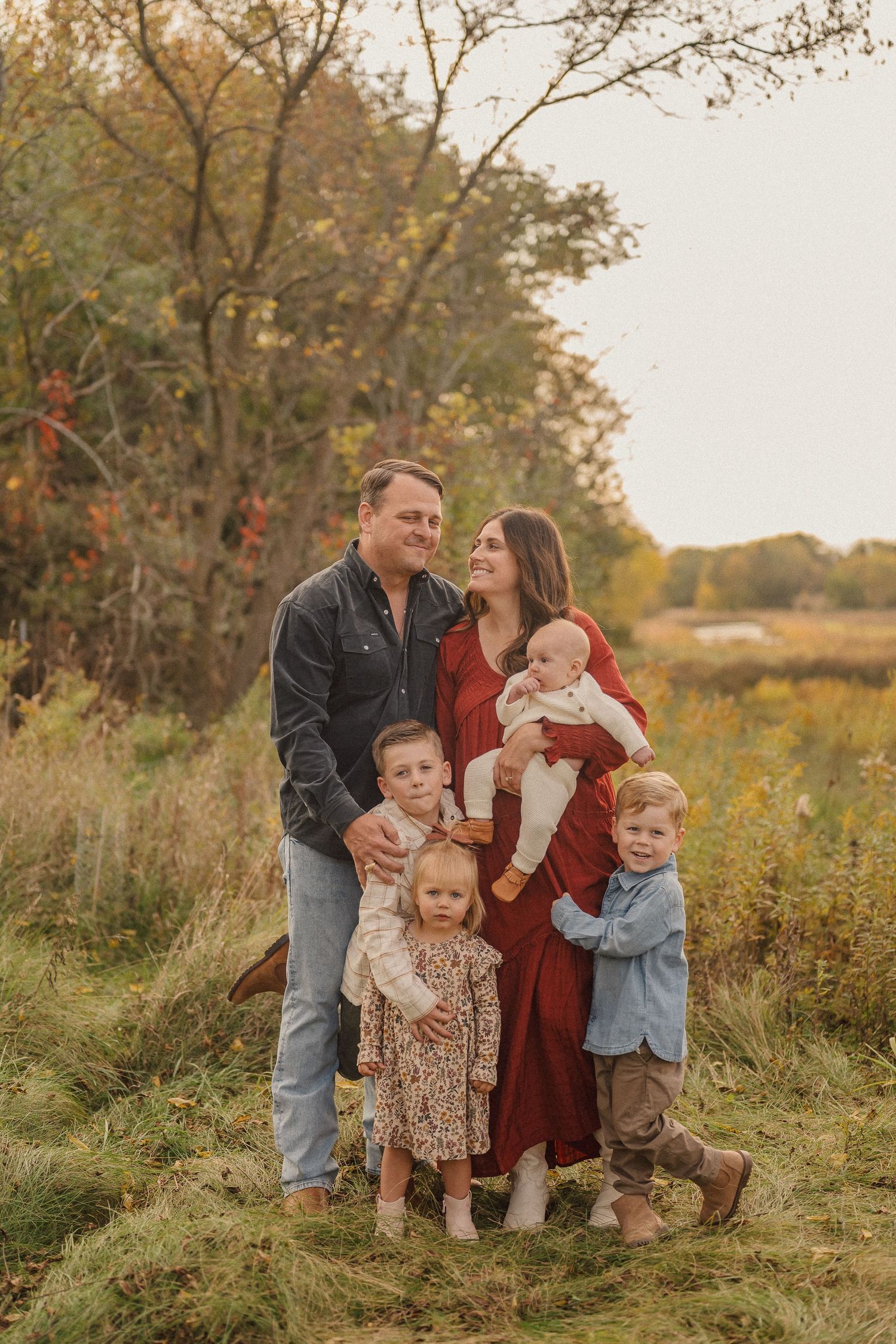 A family poses together in an autumn field at sunset wearing coordinated casual outfits.