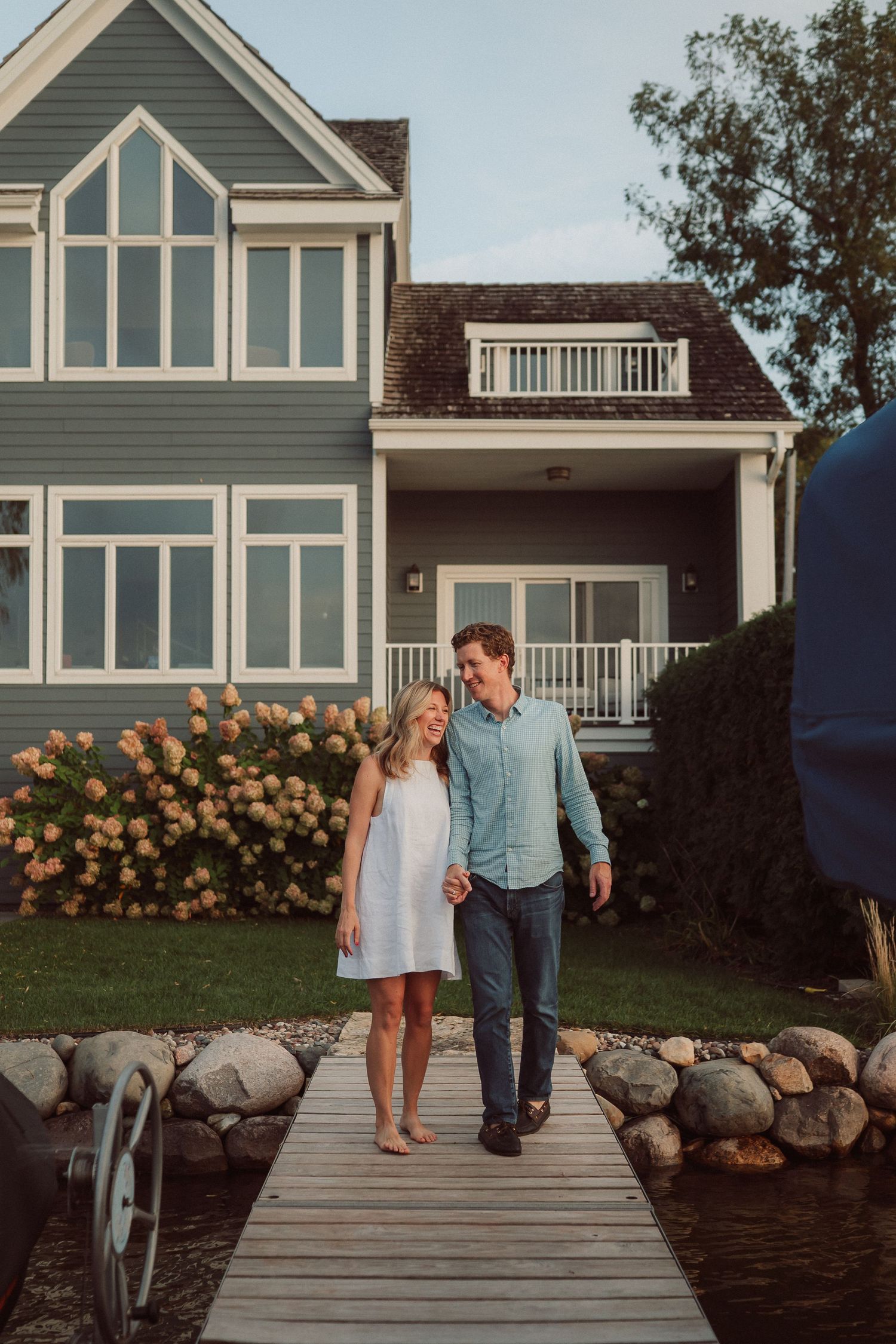A couple walks along a wooden dock with a beautiful lakeside home and hydrangea bushes in the background.