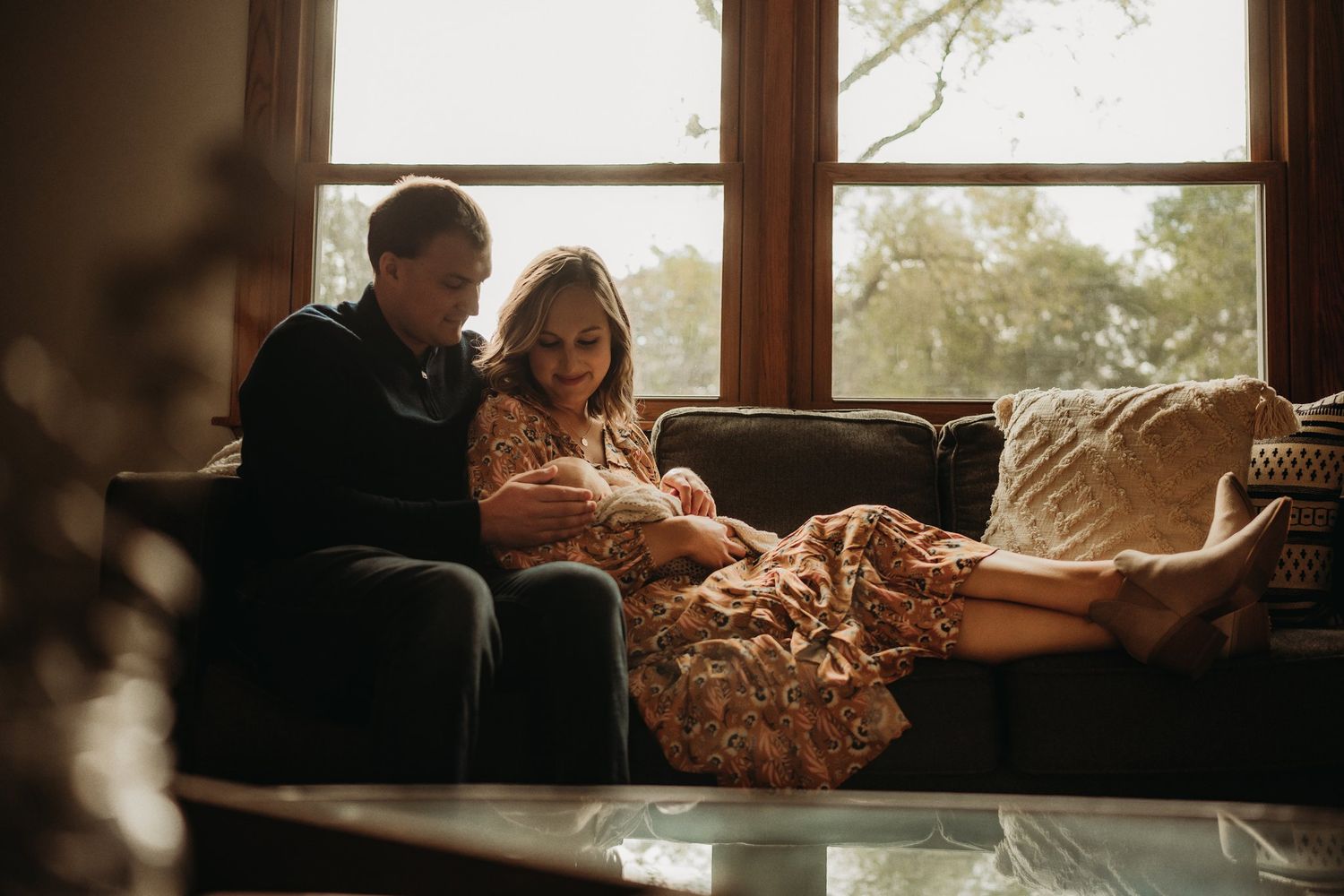 Warm sunlit photo of couple with newborn relaxing on living room couch.