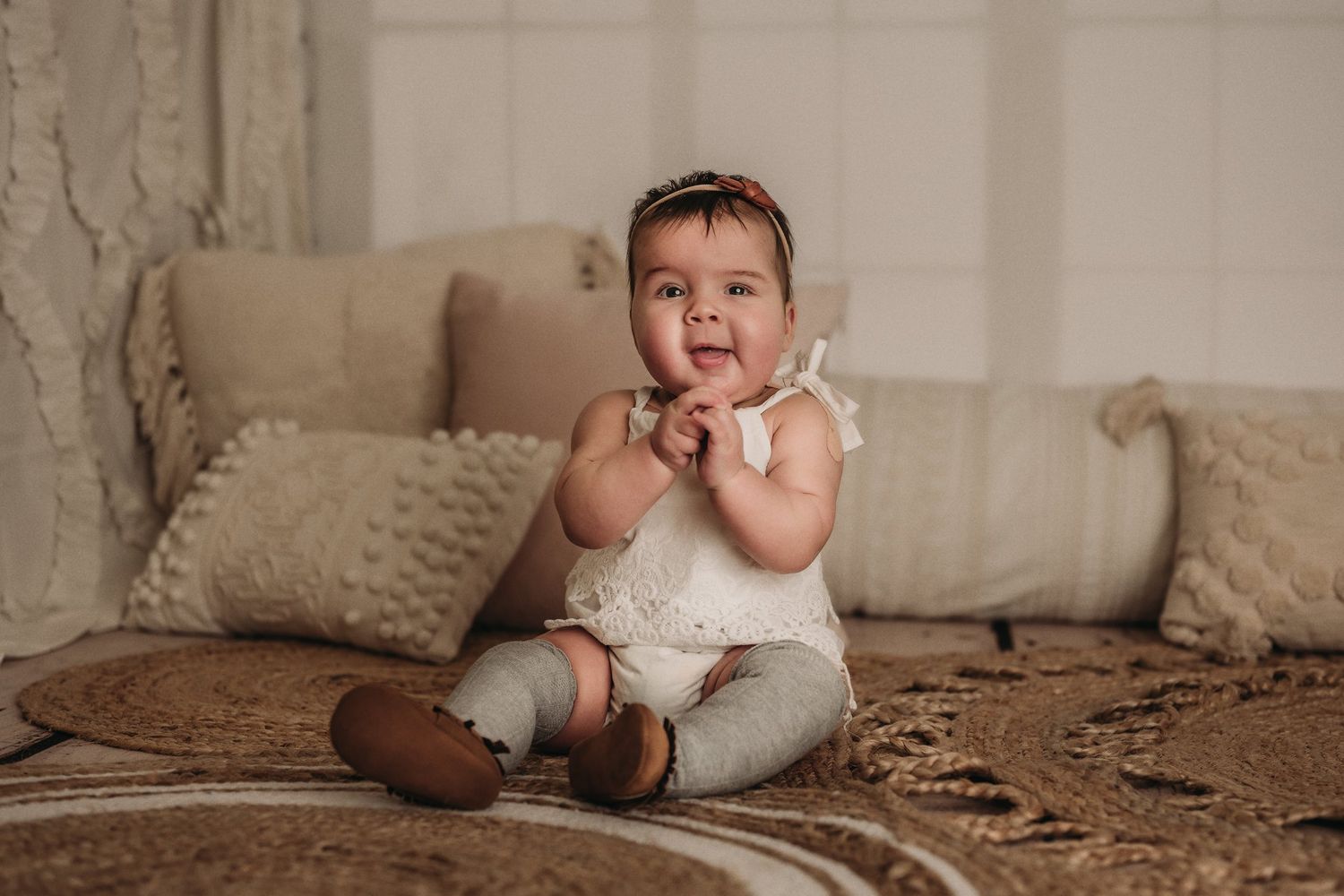 Baby in white romper poses with hands near face on textured brown carpet.