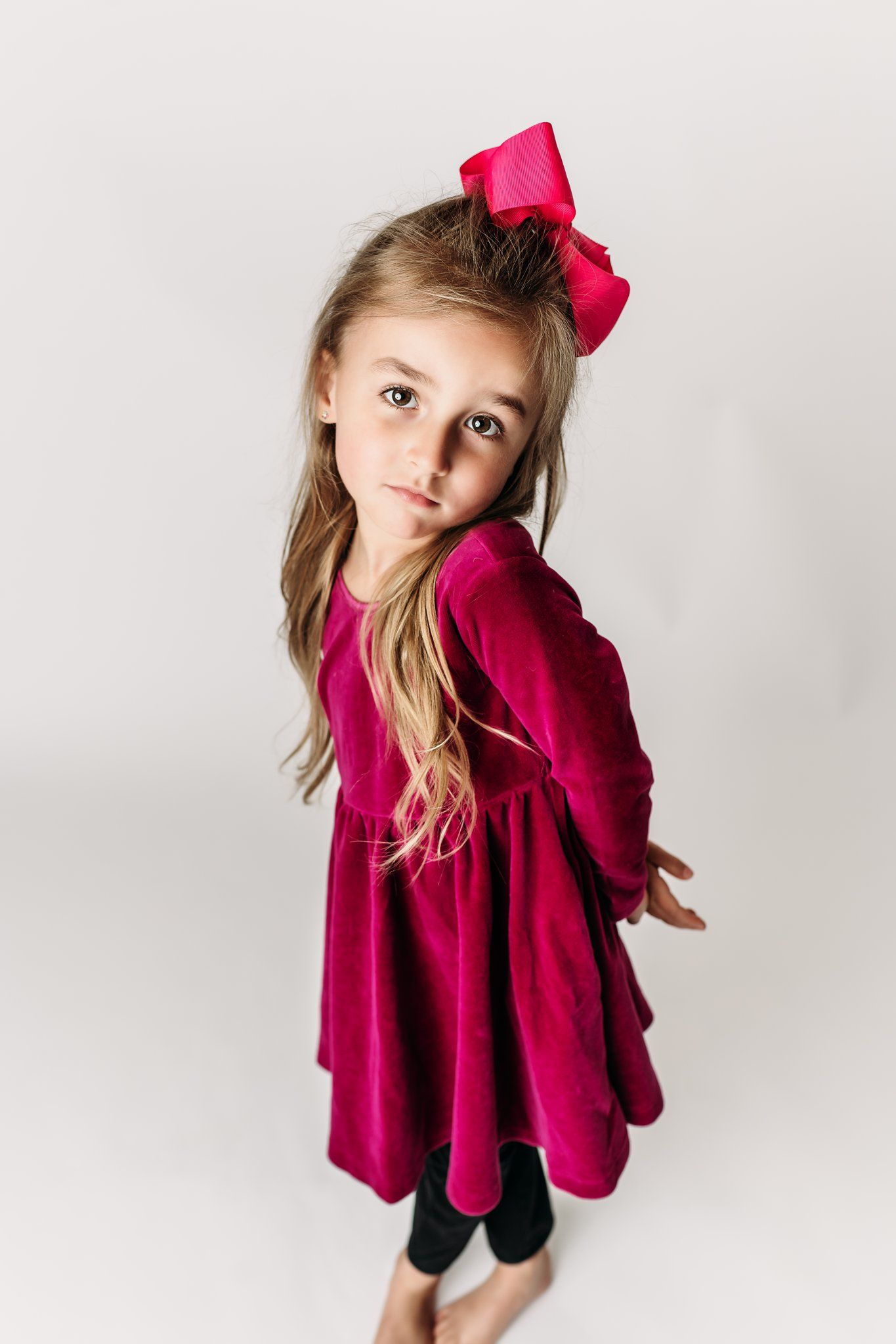 Child in velvet dress with pink bow striking whimsical pose against white background.