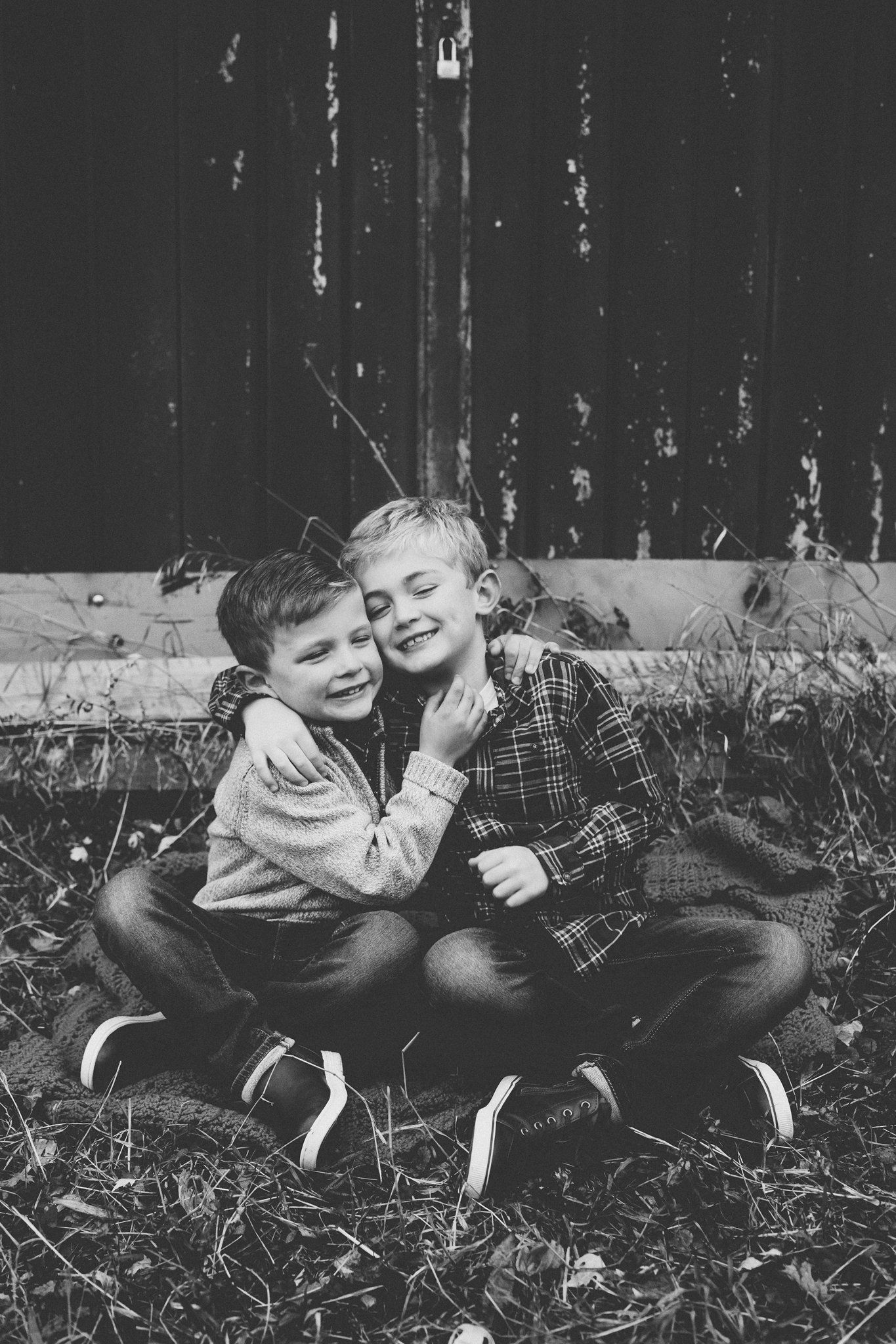 Two children share a joyful embrace while sitting together on grass in black and white.