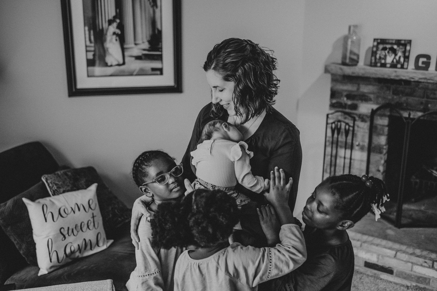 A black and white image shows a group gathered lovingly around a newborn in a living room.