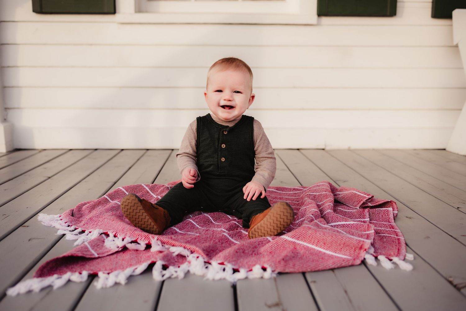 Baby sitting on pink striped blanket on wooden deck wearing black outfit.